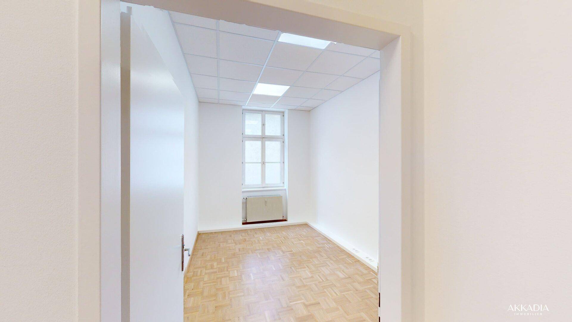 Blick durch eine offene Tür in einen hellen Raum mit Parkettboden und Fenster.