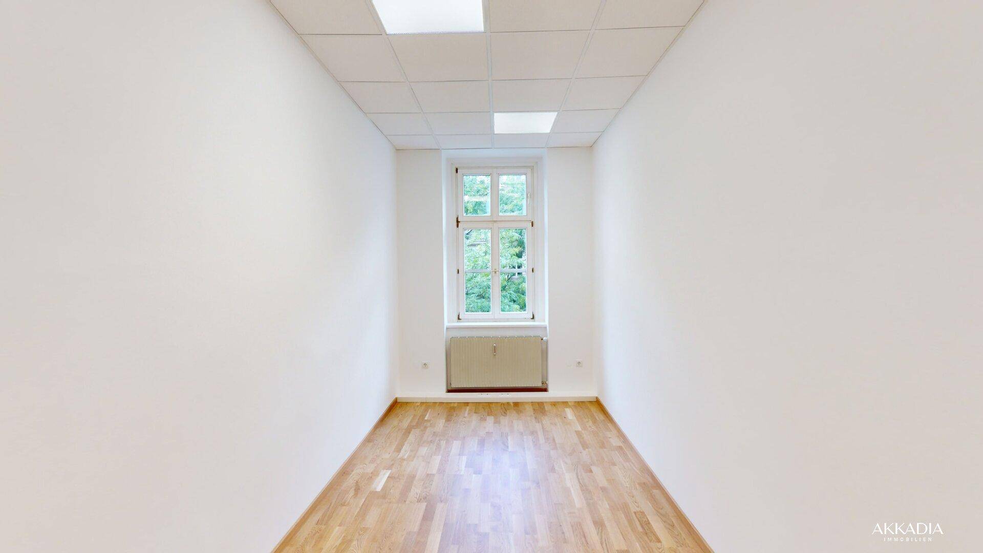 Leerer Raum mit hellem Parkettboden, weißen Wänden und einem Fenster mit Grünblick.