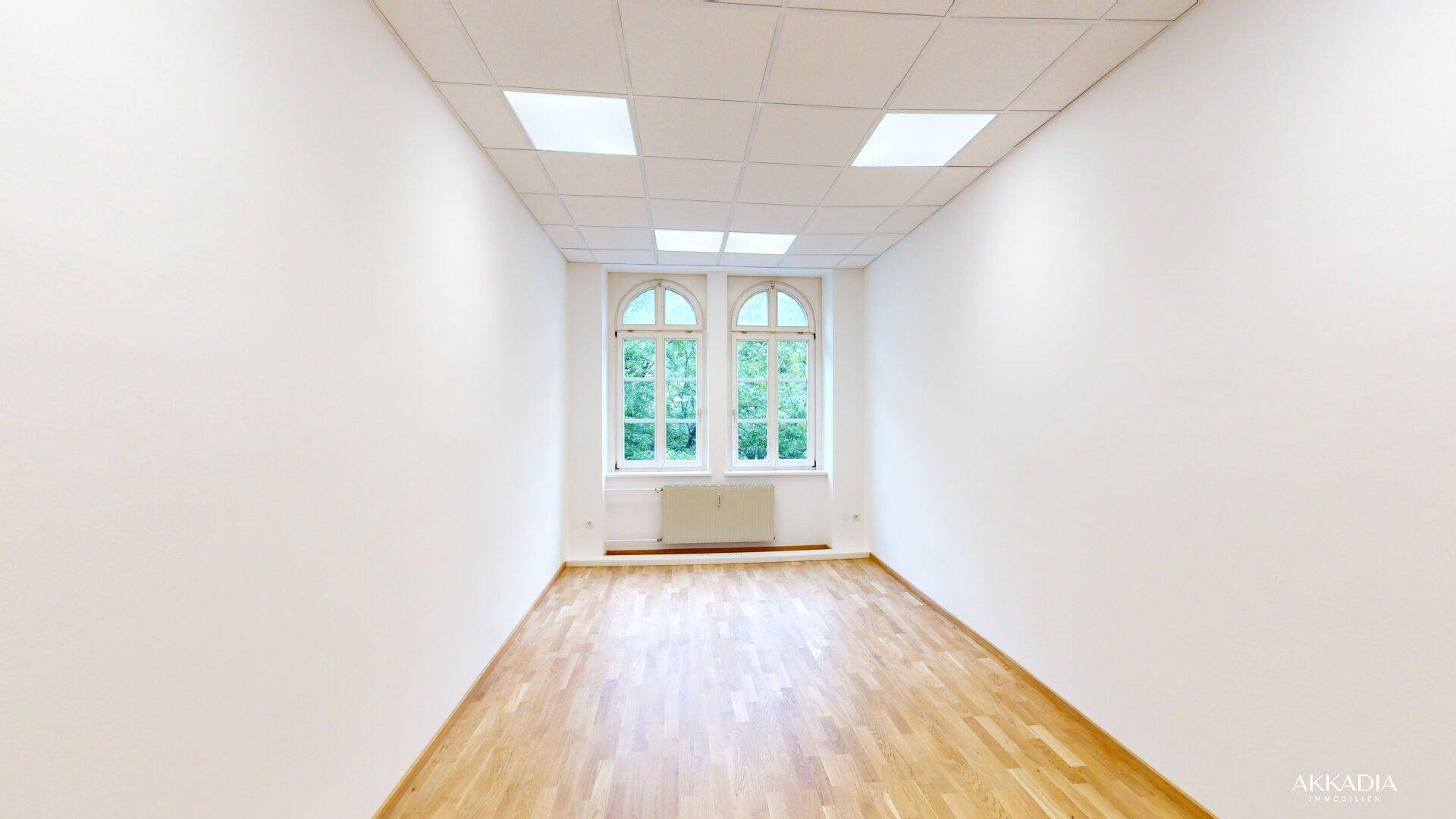 Geräumiger Raum mit Parkettboden, weißen Wänden und zwei Bogenfenstern mit Grünblick.