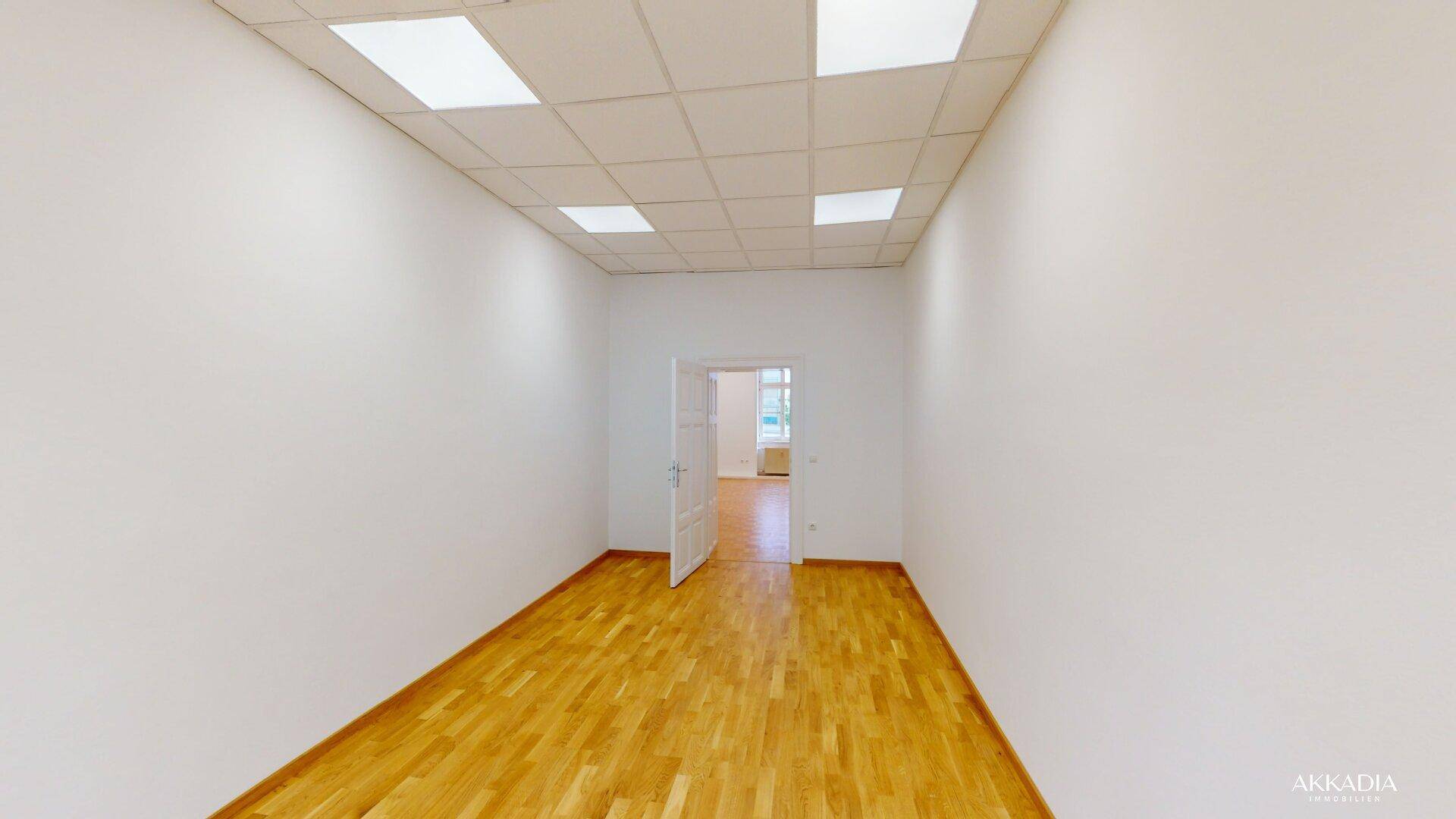 Heller, leerer Raum mit Parkettboden, weißen Wänden und offener Tür zu einem weiteren Zimmer.