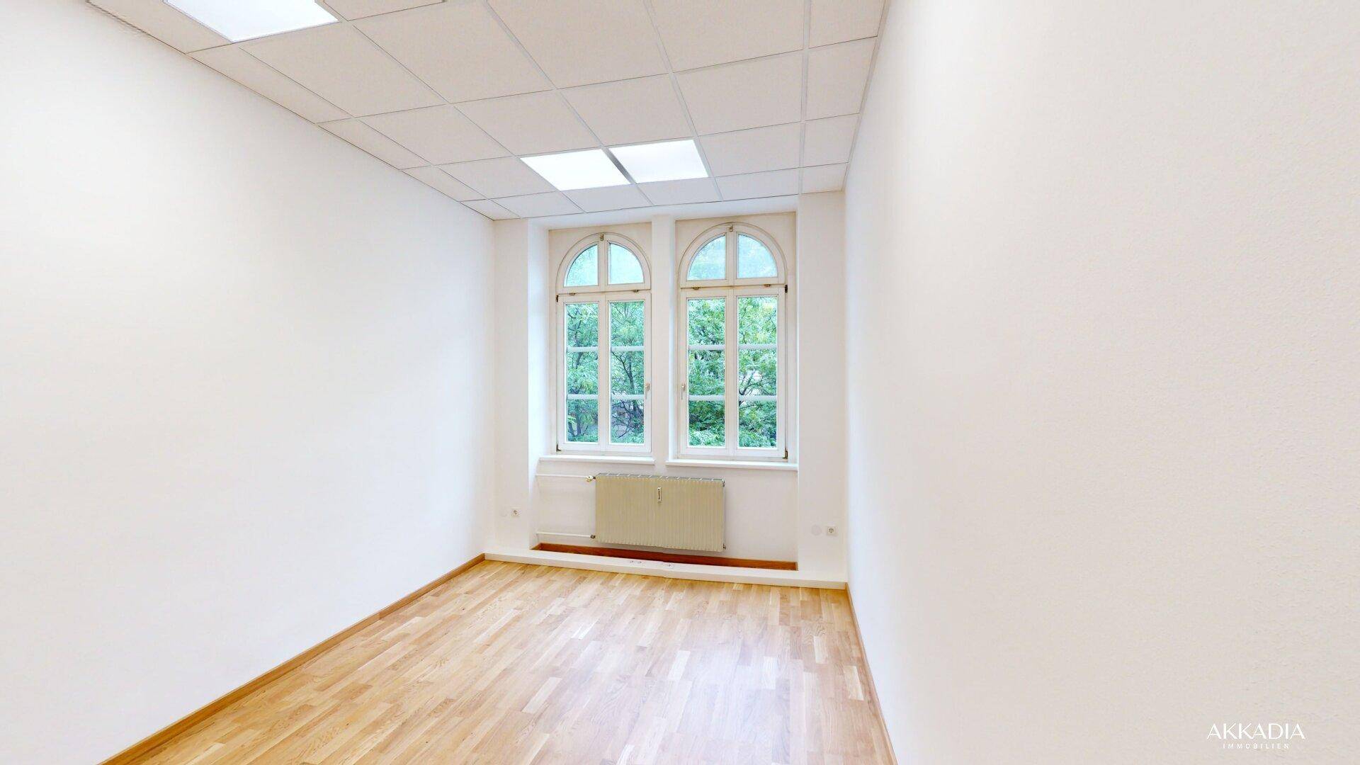 Heller Raum mit Parkettboden, weißen Wänden und zwei Bogenfenstern mit Grünblick.
