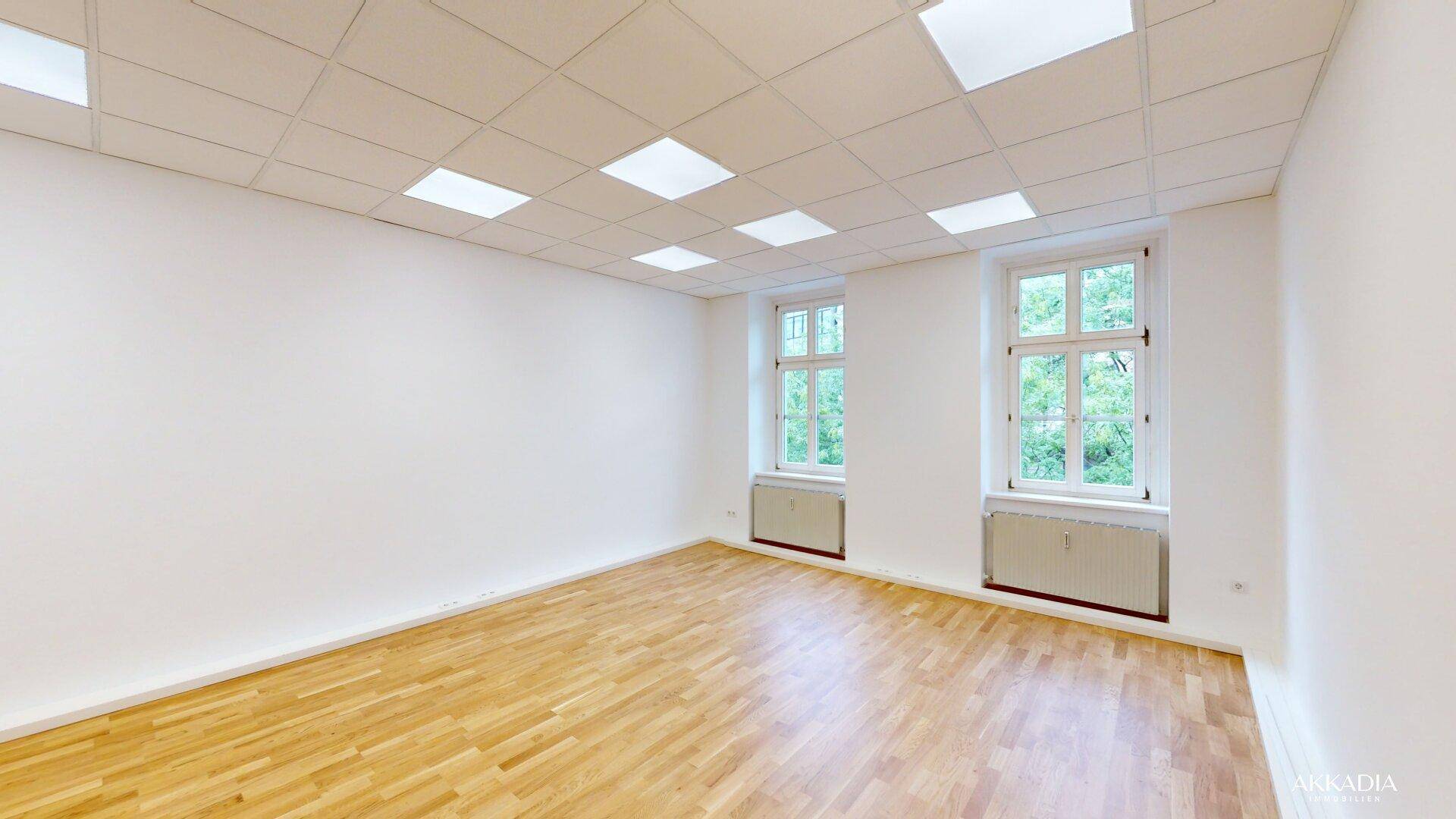 Heller, geräumiger Raum mit Parkettboden, weißen Wänden und zwei Fenstern mit Grünblick.