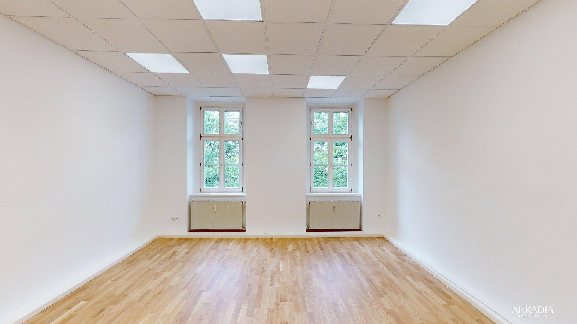 Geräumiger, heller Raum mit Parkettboden, weißen Wänden und zwei Fenstern mit Grünblick.