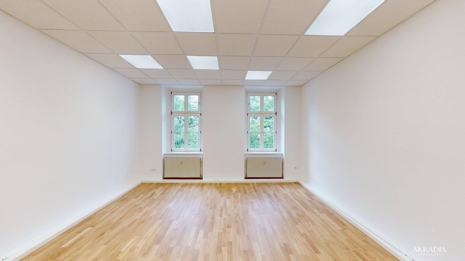 Geräumiger, heller Raum mit Parkettboden, weißen Wänden und zwei Fenstern mit Grünblick.