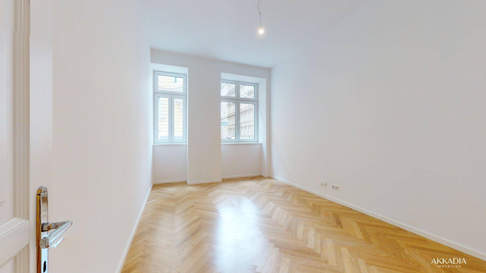 Lichtdurchflutetes Zimmer mit zwei großen Fenstern und elegantem Fischgrätparkett.