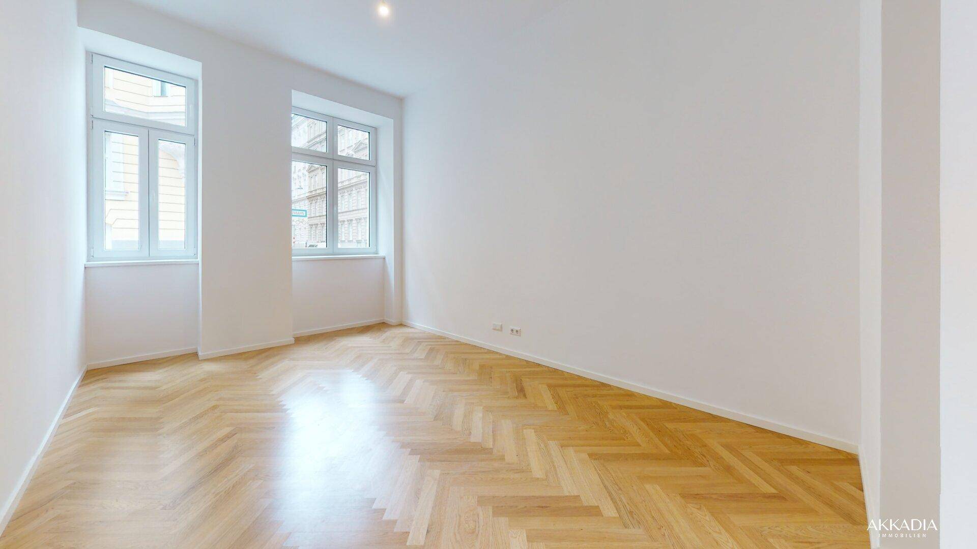 Lichtdurchflutetes Zimmer mit zwei großen Fenstern und elegantem Fischgrätparkett.
