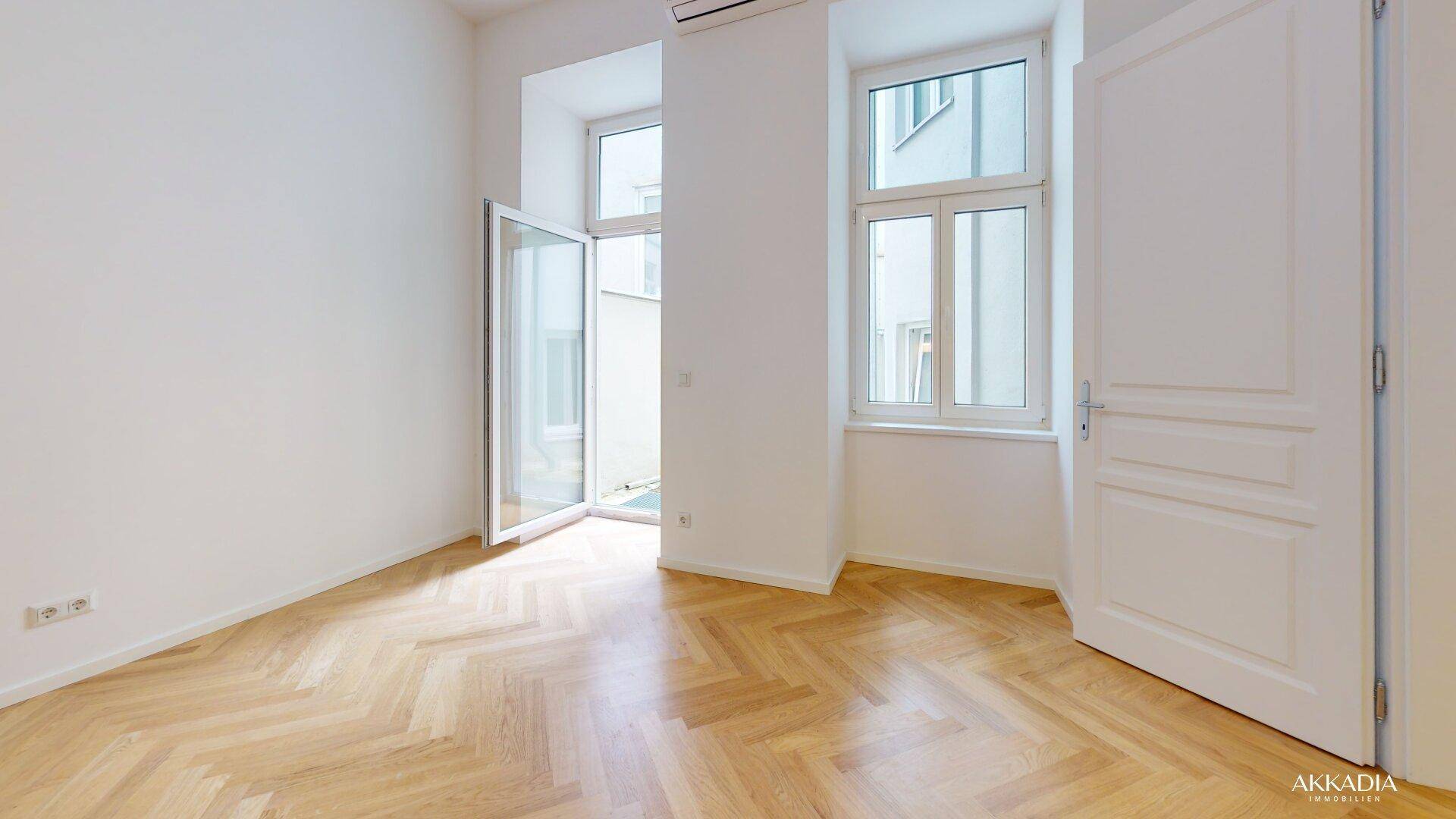 Helles Zimmer mit Fischgrätparkett, Fenster und Zugang zu einem kleinen Innenhof.
