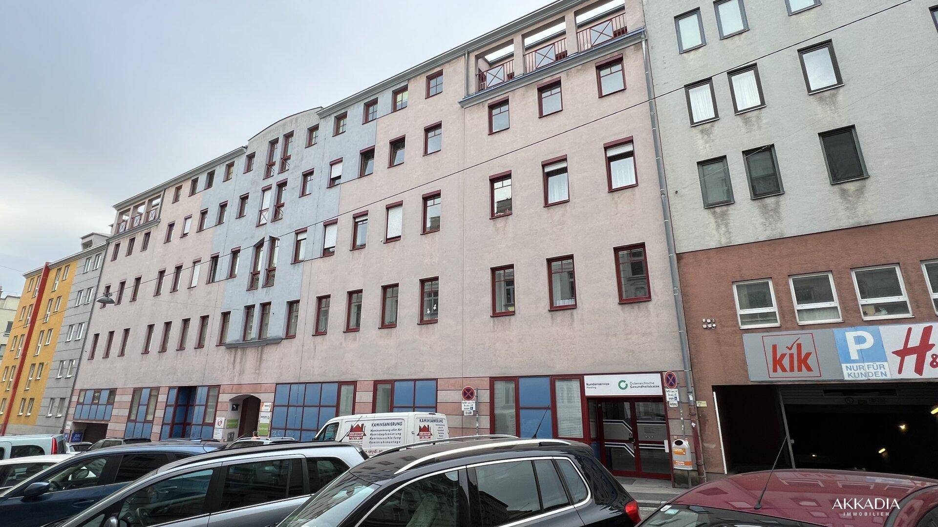 Mehrstöckiges Gebäude mit vielen Fenstern und Geschäften im Erdgeschoss, umgeben von parkenden Autos.