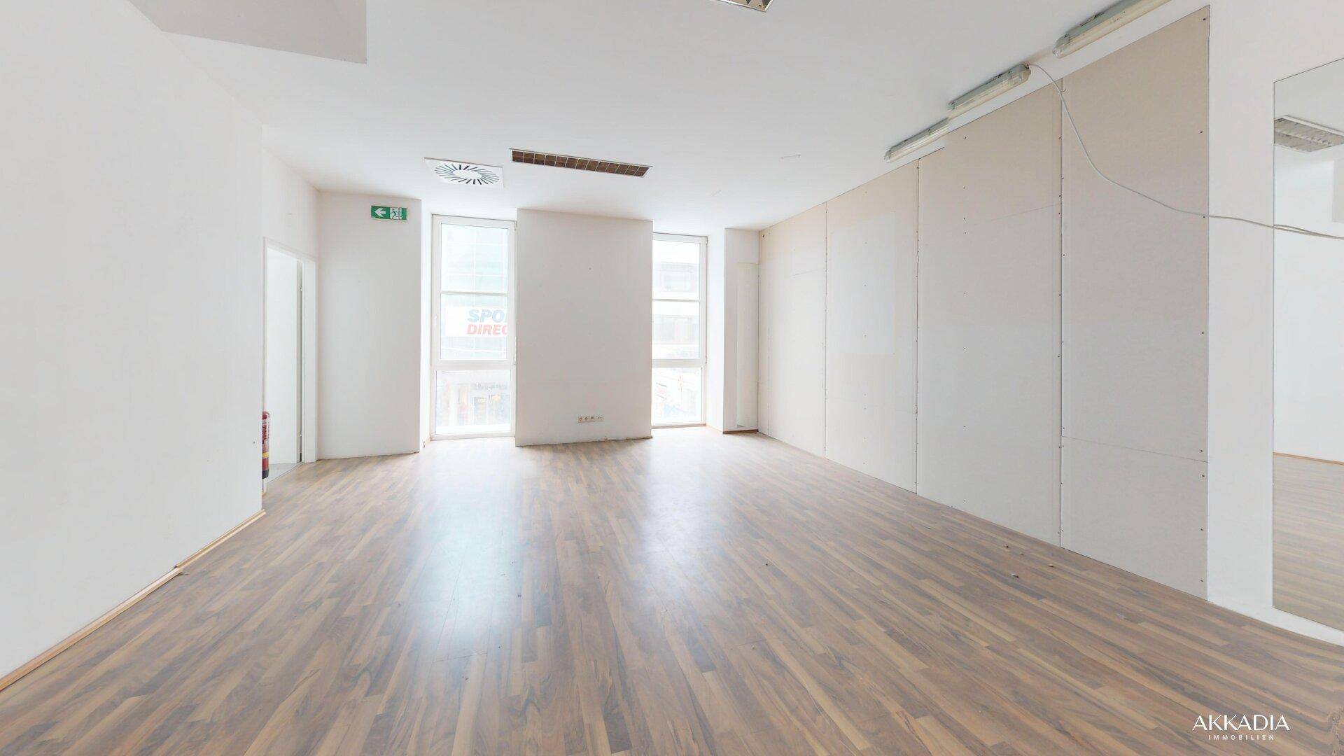 Geräumiger Büroraum mit Laminatboden und hohen Fenstern, bietet viel Tageslicht und Platz.