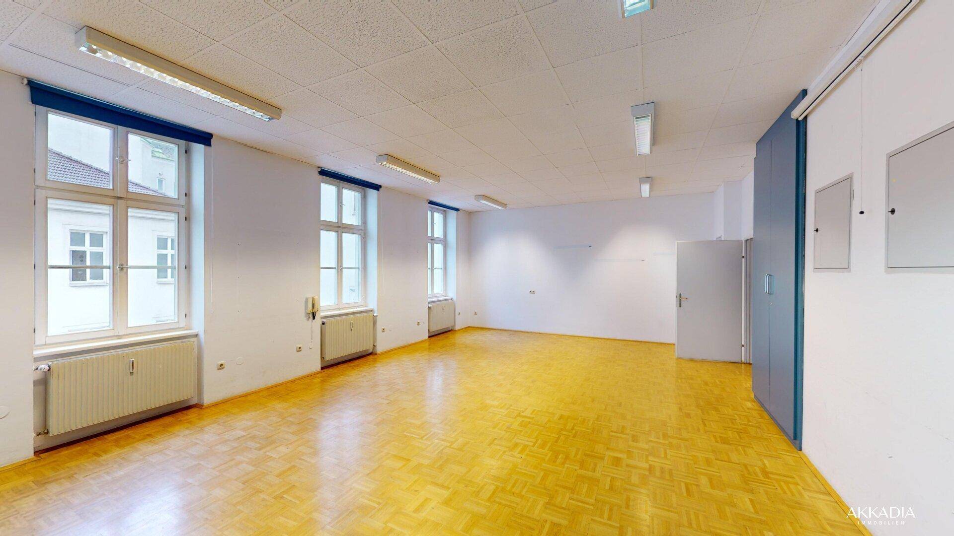 Heller Büroraum mit klassischem Parkettboden, hohen Fenstern und Heizkörpern unter den Fensterbänken.