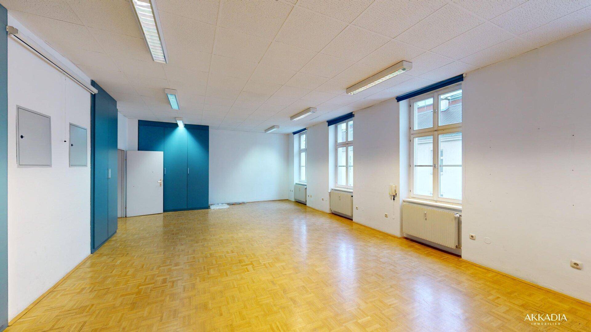 Geräumiger Büroraum mit Parkettboden, weißen Wänden und blauen Einbauschränken, bietet viel Platz.