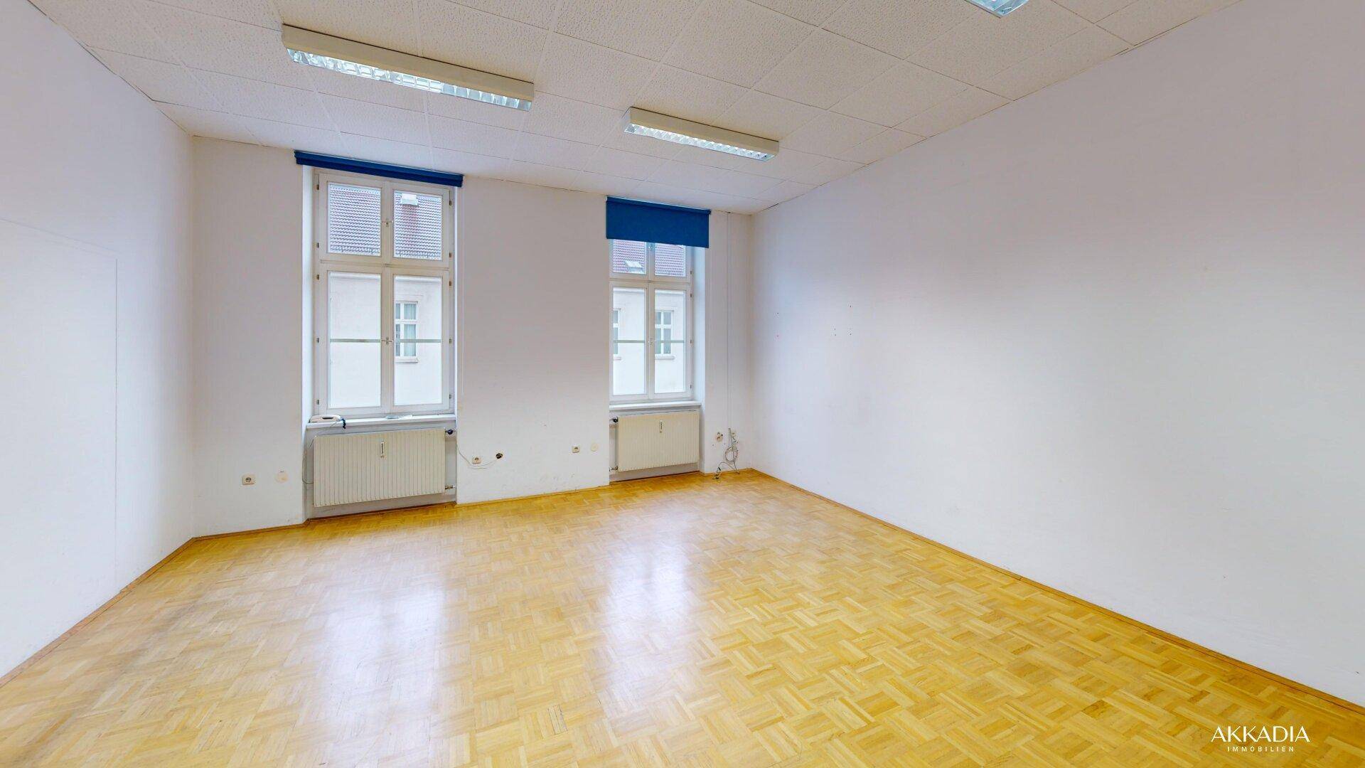Heller Büroraum mit Parkettboden und zwei großen Fenstern, die viel Tageslicht hereinlassen.