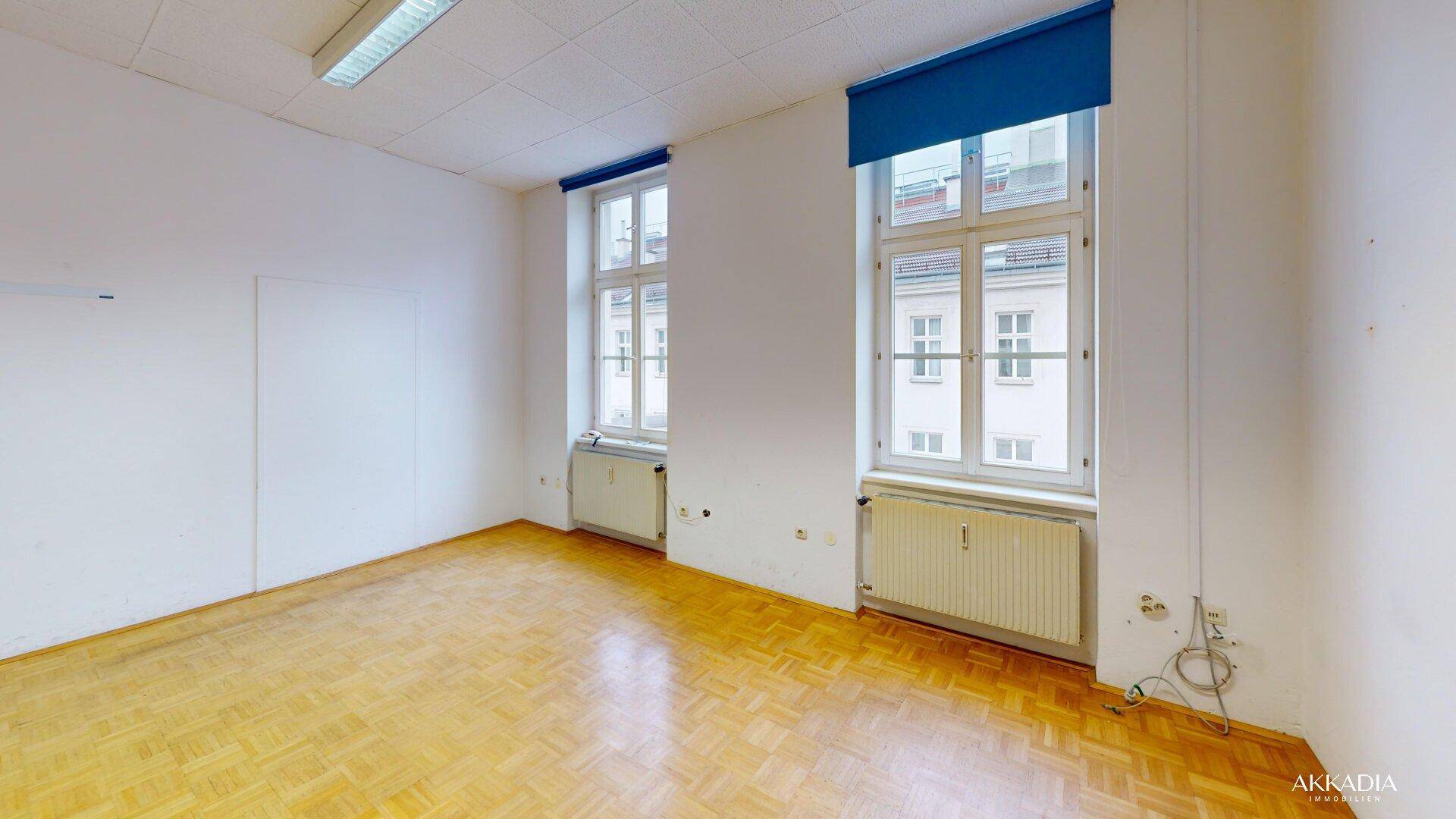 Heller Büroraum mit Parkettboden und zwei großen Fenstern, die viel Tageslicht hereinlassen.