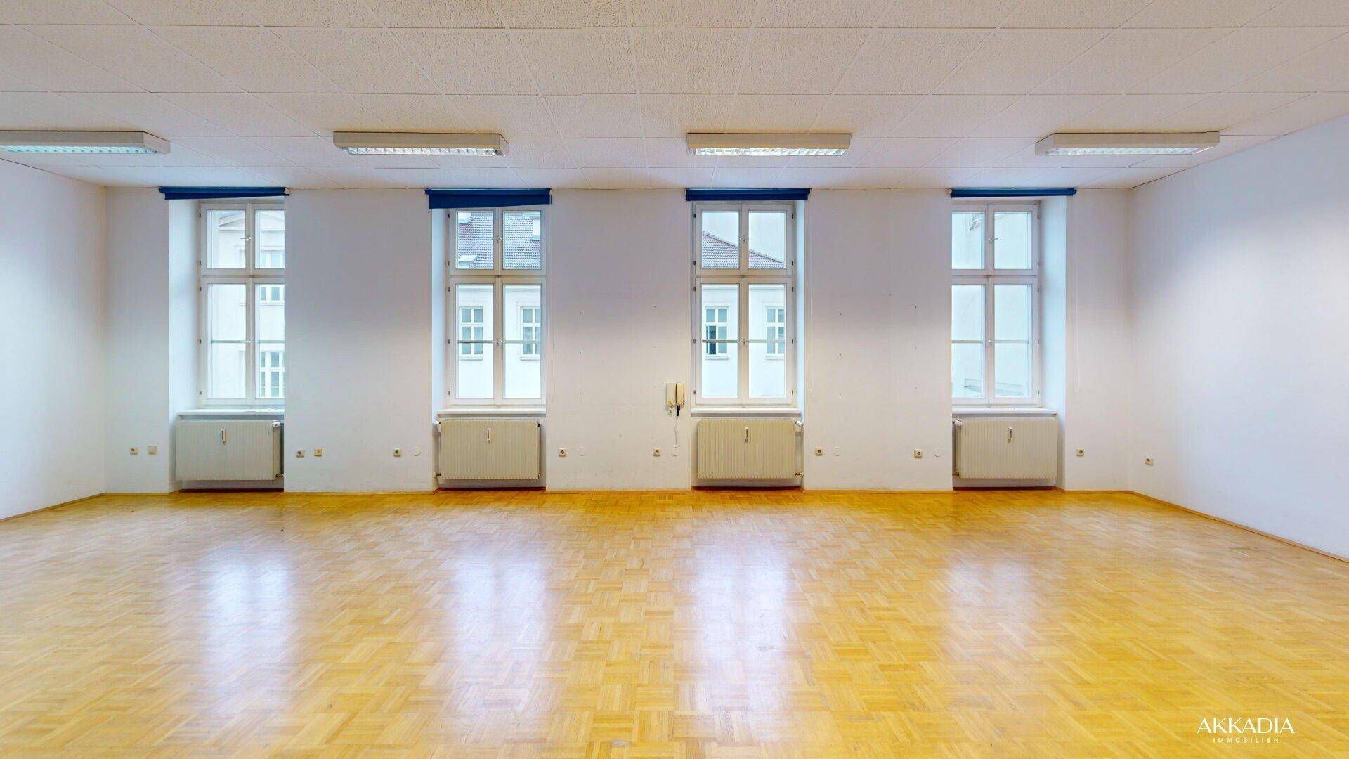 Geräumiger, heller Raum mit Fischgrätparkett, weißen Wänden und mehreren Fenstern, ideal für Büro oder Wohnen.