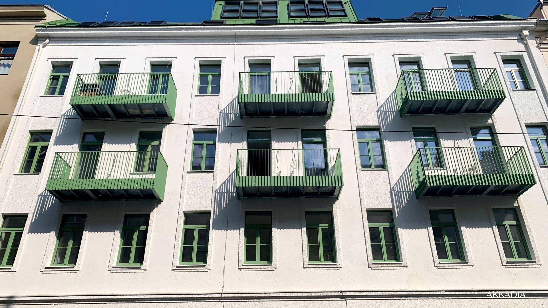 Weiße Fassade eines Mehrfamilienhauses mit grünen Balkonen und Fenstern unter blauem Himmel.