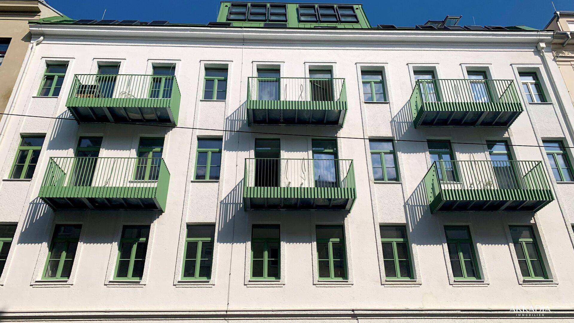 Frontansicht eines Gebäudes mit symmetrisch angeordneten grünen Balkonen und vielen Fenstern.