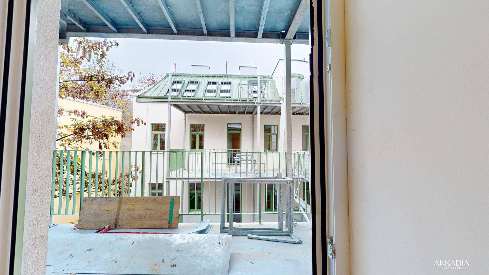 Unfertiger Balkon mit Blick auf ein weiteres Gebäude, umgeben von Bäumen und einem grünen Zaun.