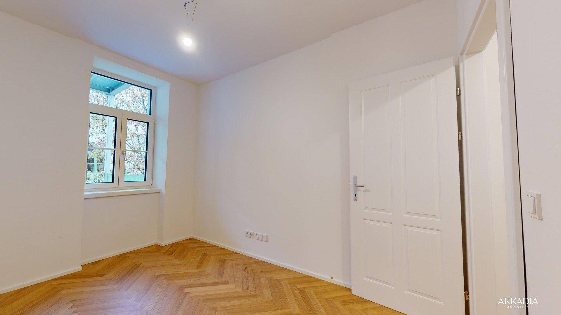 Leeres Zimmer mit Fischgrätparkett und einem Fenster, das viel Tageslicht hereinlässt.