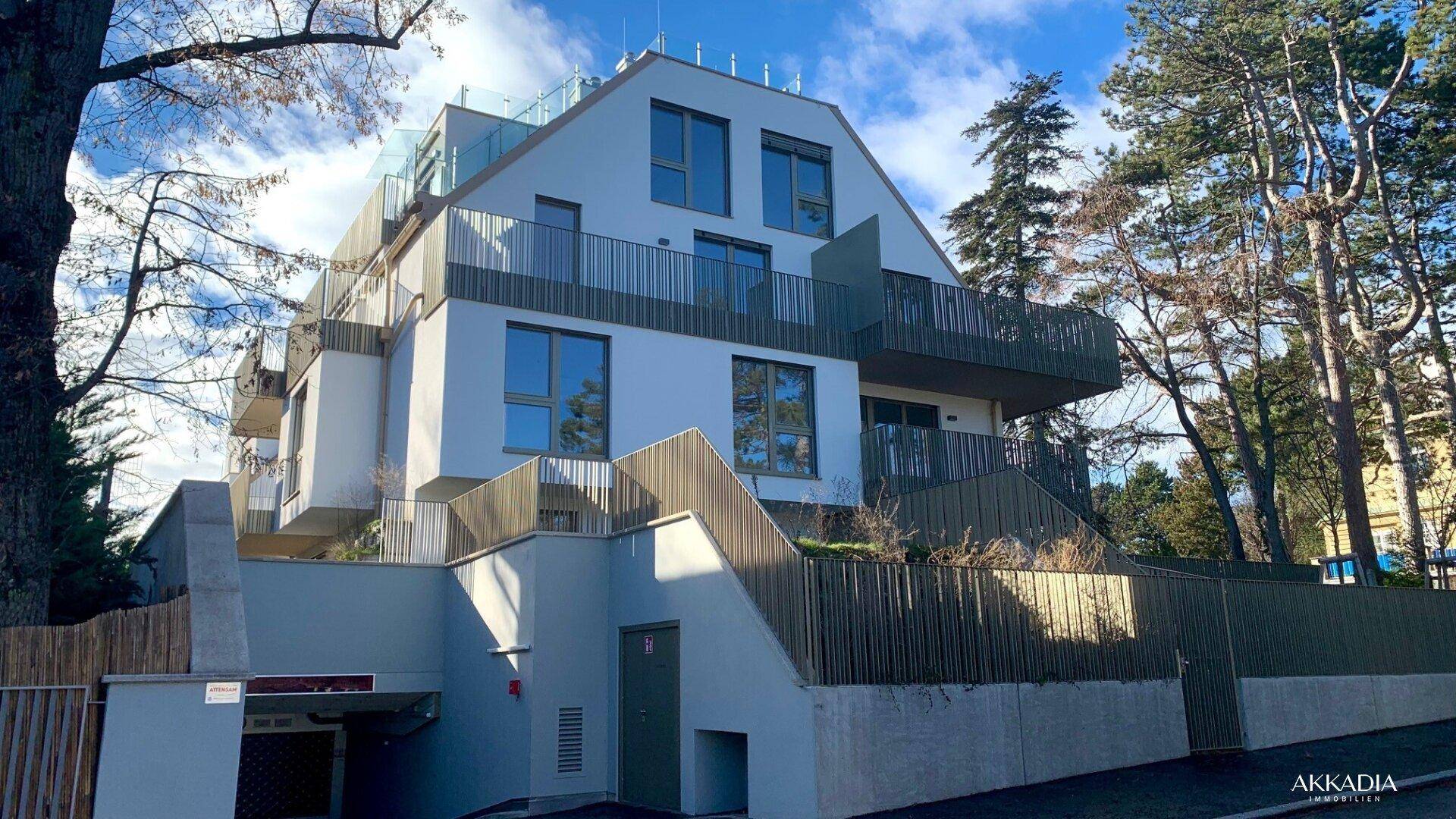 Modernes Wohngebäude mit mehreren Etagen, Balkonen und Tiefgaragenzufahrt, umgeben von Bäumen.