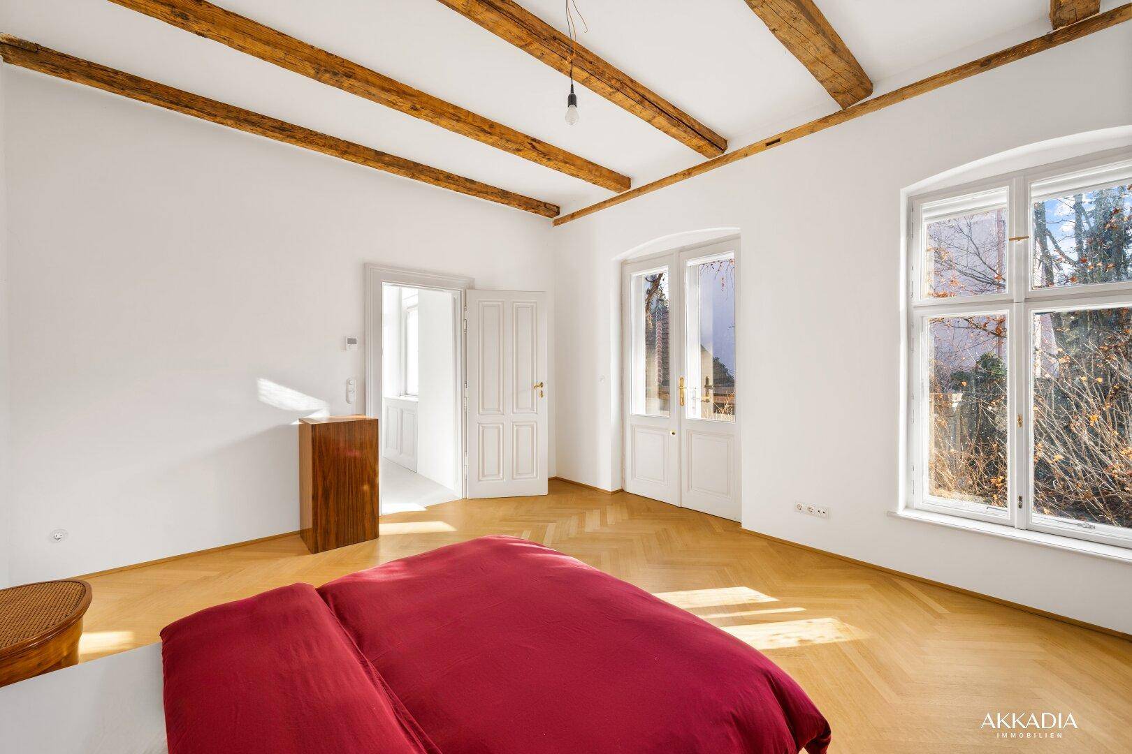 Helles Schlafzimmer mit Fischgrätparkett, freiliegenden Holzbalken und Zugang zu einem Balkon.