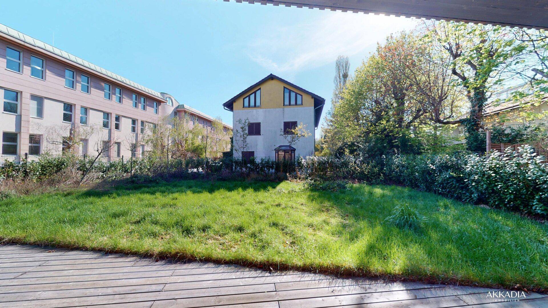 Blick von der Holzterrasse auf eine grüne Rasenfläche und umliegende Gebäude bei sonnigem Wetter.