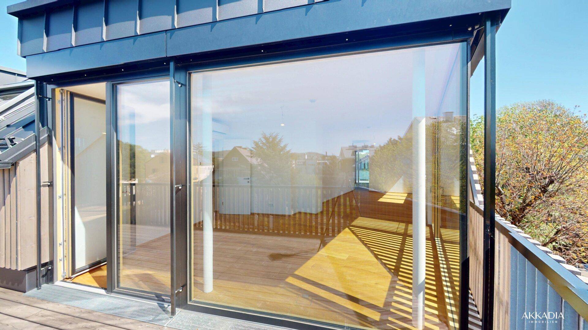 Sonniger Terrassenbereich mit Glasgeländer und Blick in den hellen Innenraum.