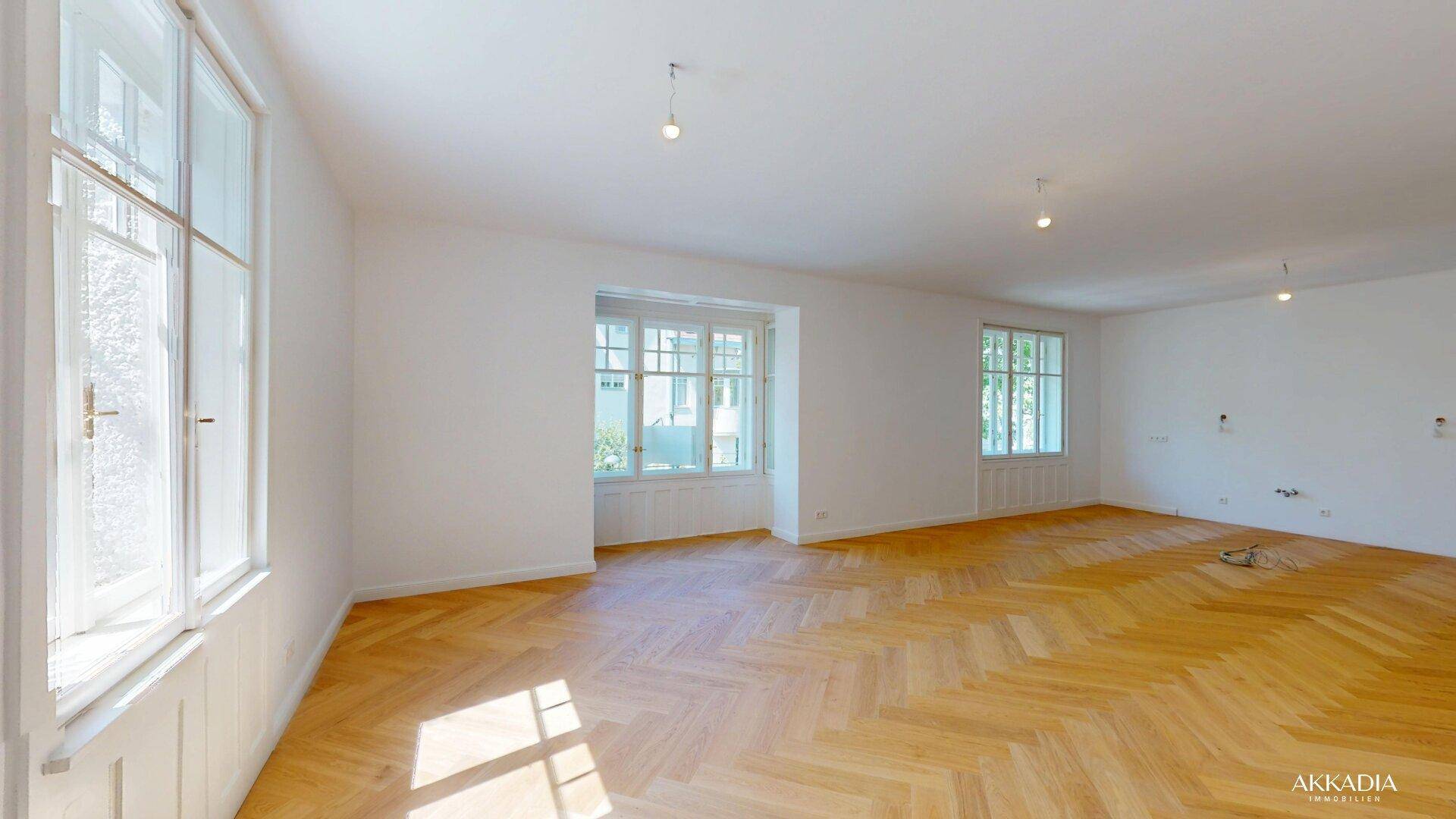 Großes, lichtdurchflutetes Zimmer mit Fischgrätparkett und mehreren Fenstern.