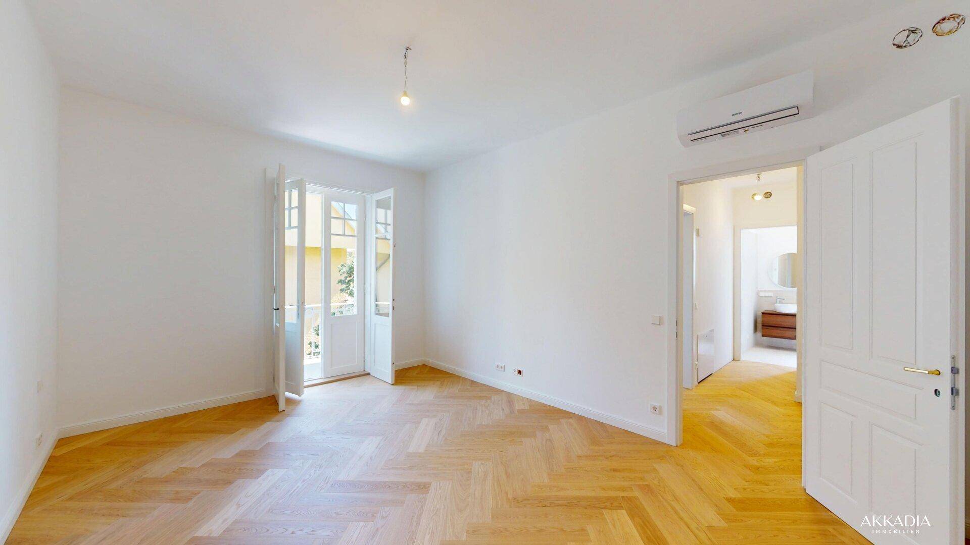 Helles Zimmer mit Fischgrätparkett und Zugang zu einem kleinen Balkon.