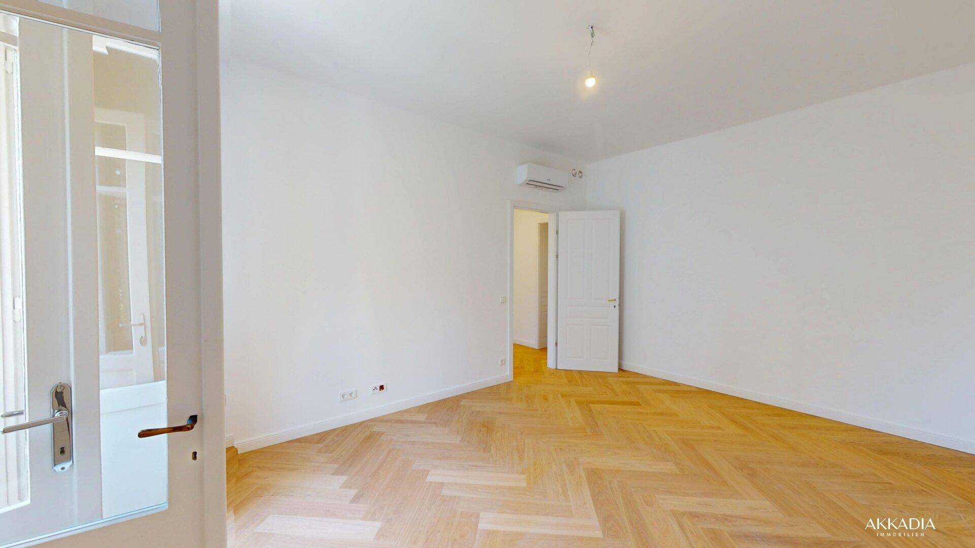 Helles Zimmer mit Fischgrätparkett, Klimaanlage und Zugang zu einem Balkon.