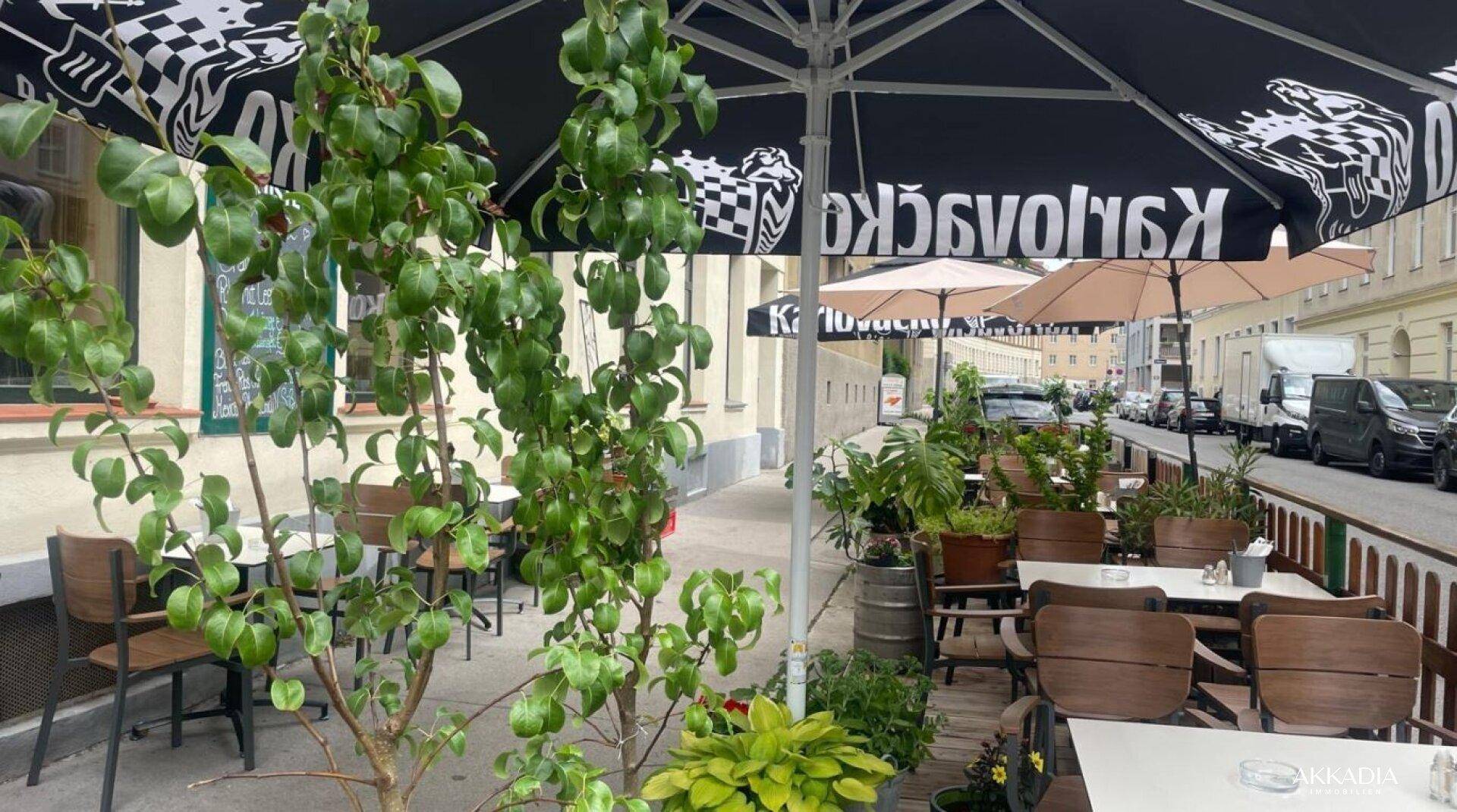Eine Außenterrasse mit Tischen, Stühlen und großen Sonnenschirmen, umgeben von Pflanzen.