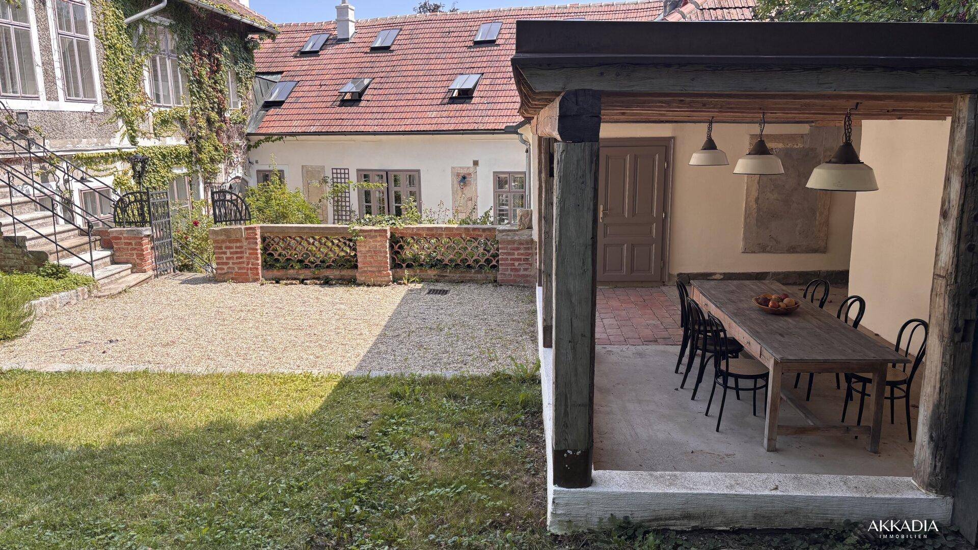 Überdachter Essbereich im Freien mit Holztisch und Stühlen, angrenzend an einen Gartenbereich.