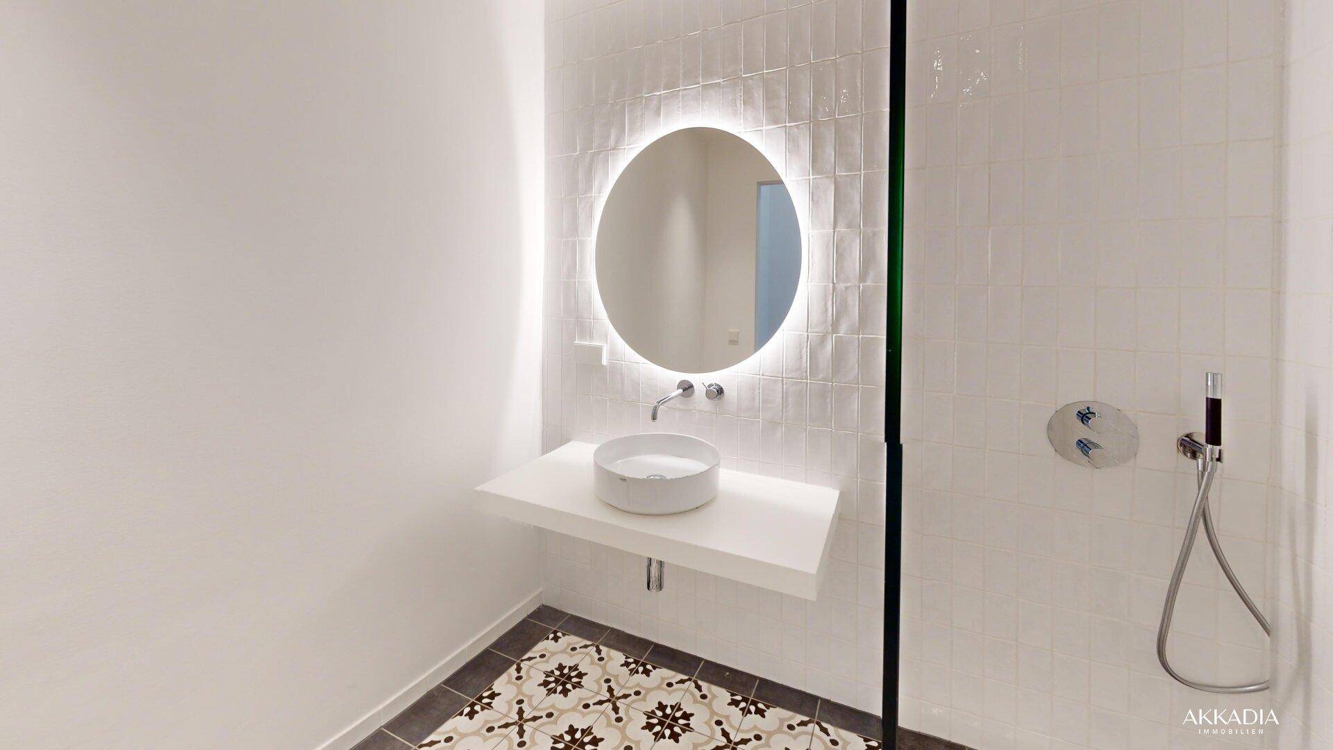 Modernes Badezimmer mit runder beleuchteter Spiegelfläche, Aufsatzwaschbecken und Duschbereich.