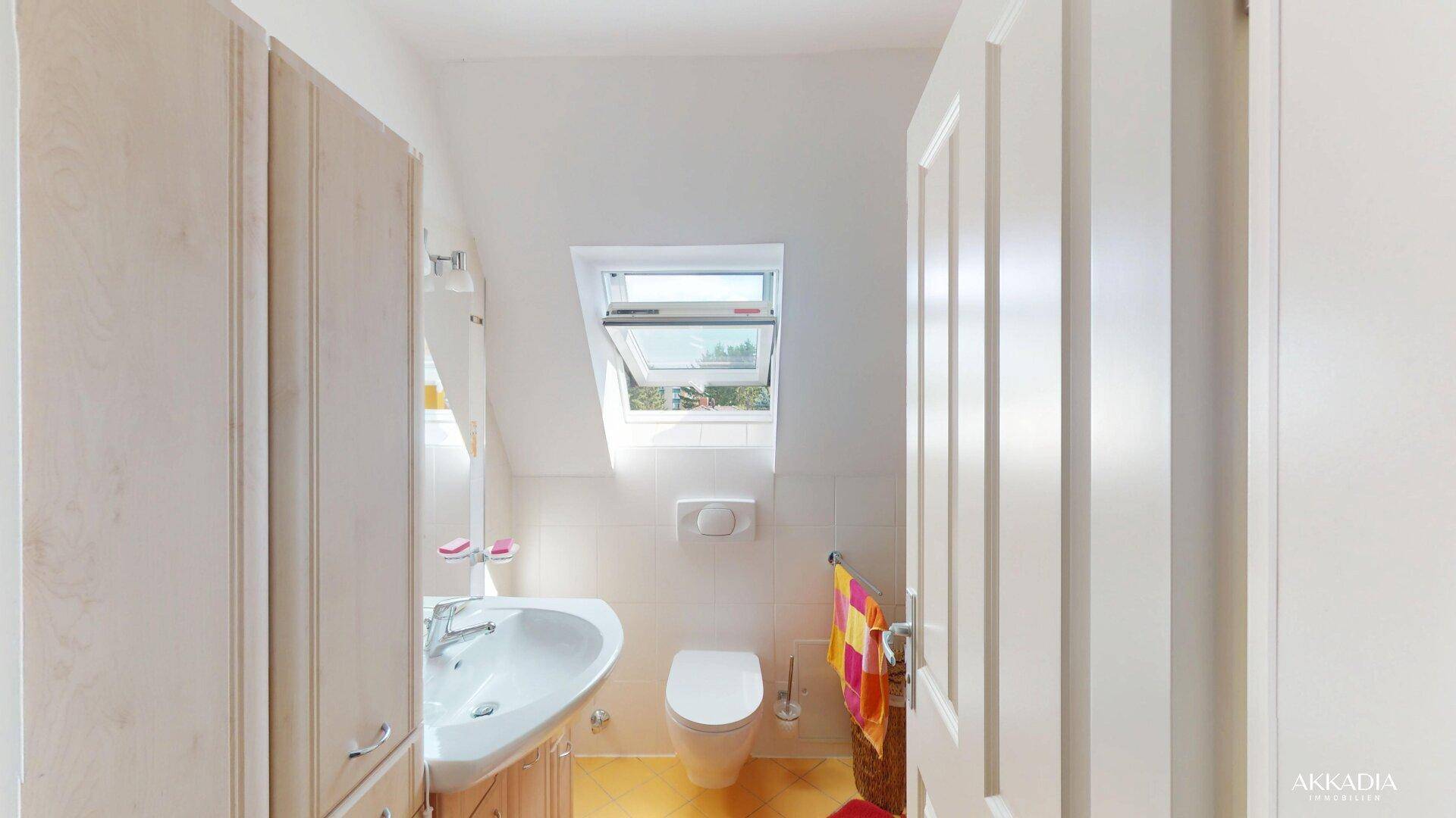 Helles Badezimmer mit Waschbecken, Toilette und Dachfenster, unter einer schrägen Decke gelegen.