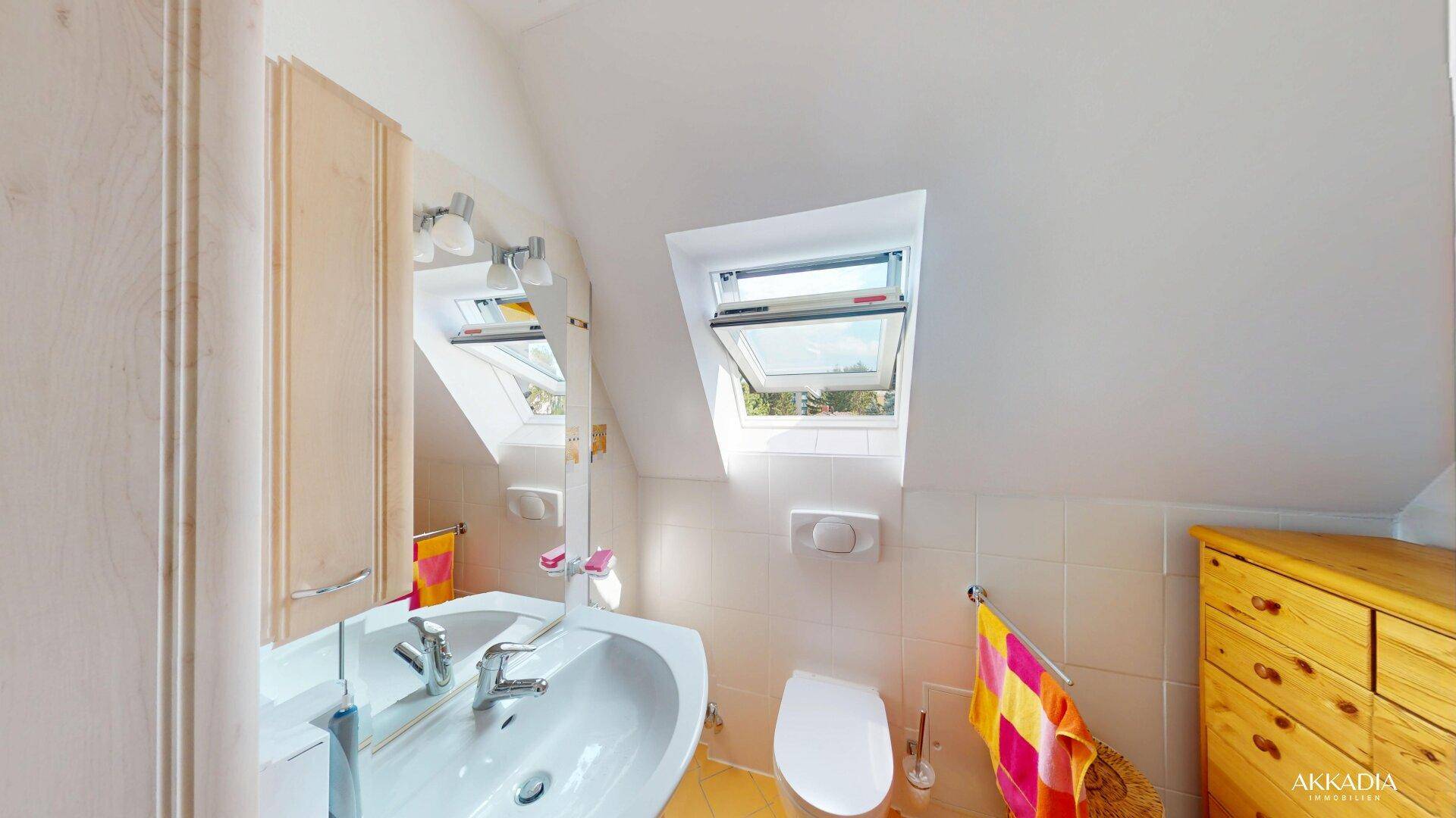 Modernes Badezimmer mit Waschbecken, Spiegel, Toilette und Dachfenster, das viel Licht hereinlässt.