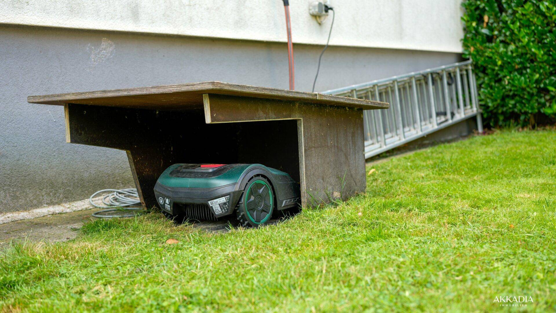 Rasenroboter unter einem Holzunterstand im Garten, mit einer Leiter an der Hauswand.