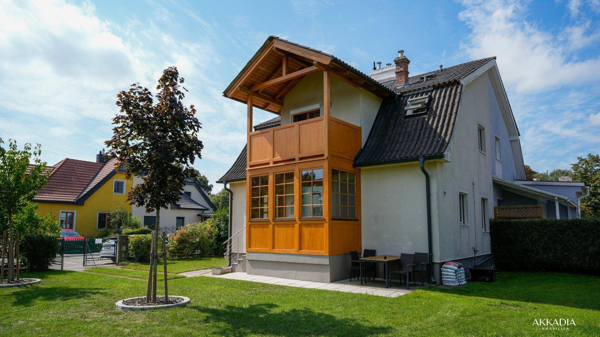 Rückansicht des Hauses mit Garten, Terrasse und traditionellem Holzbalkon unter strahlendem Himmel.