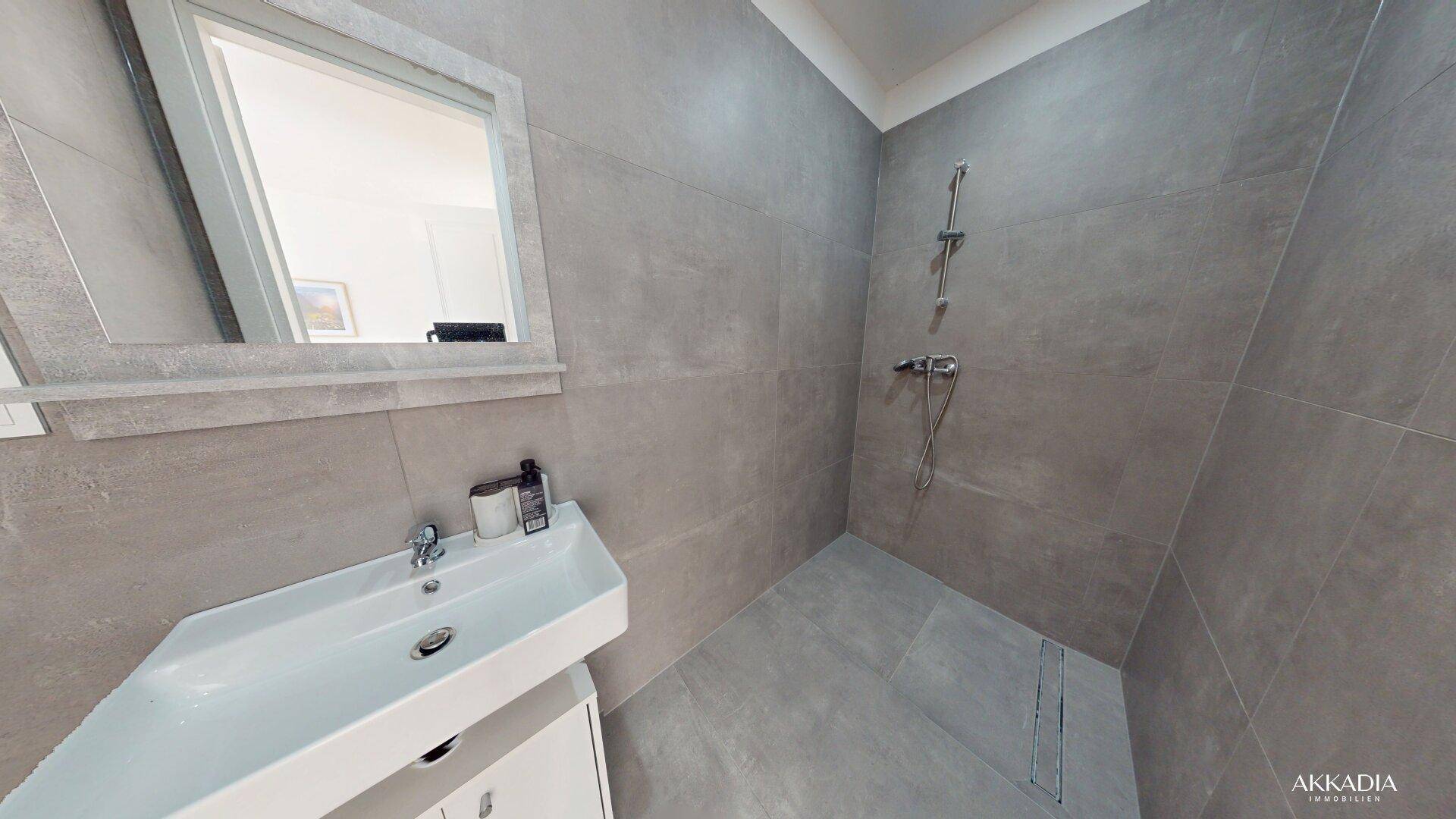 Geräumiges Badezimmer mit großen grauen Fliesen, modernem Waschbecken und begehbarer Dusche.