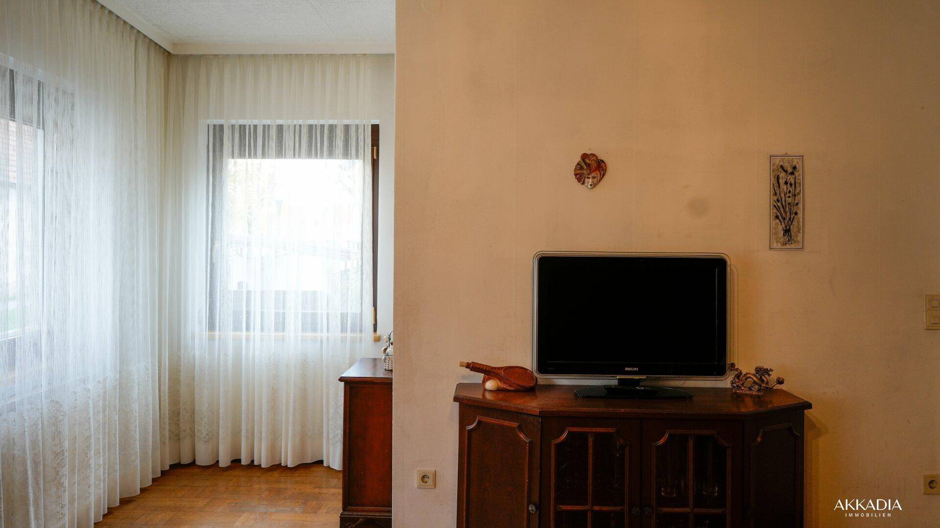 Helles Wohnzimmer mit Fernseher, Holzmöbeln und großen Fenstern.