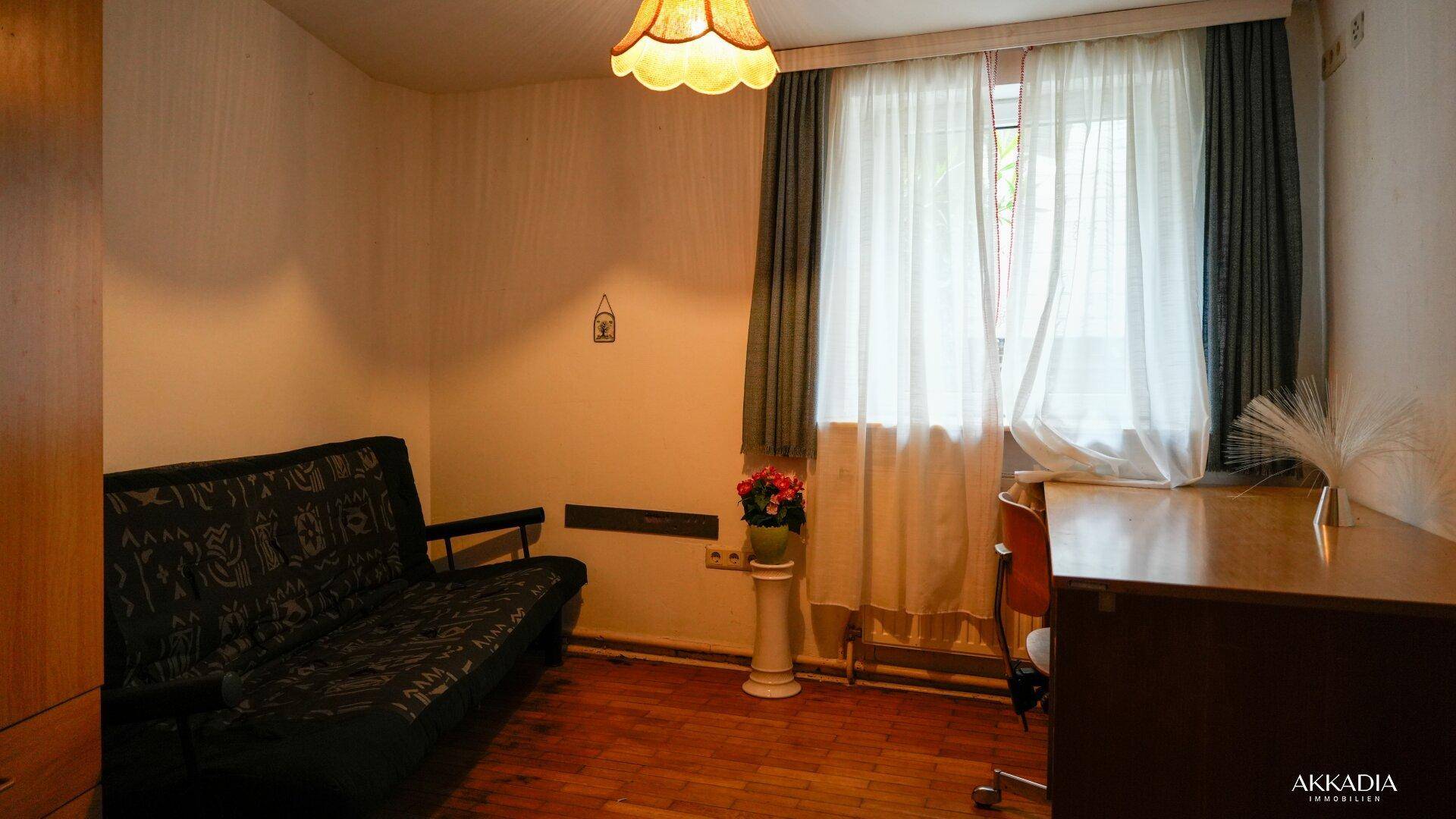 Zimmer mit Schlafsofa, Schreibtisch und Fenster mit Vorhängen.