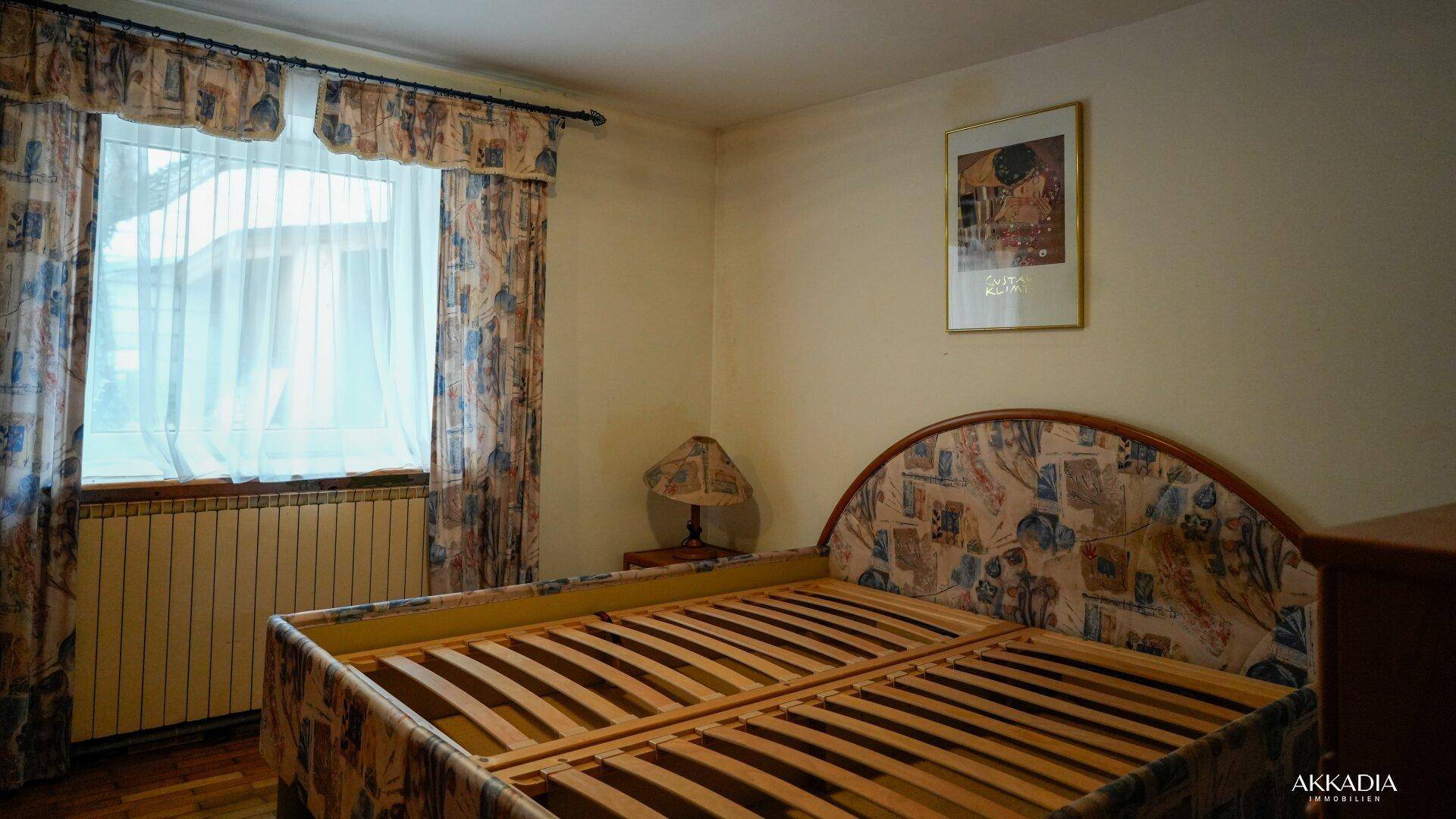 Schlafzimmer mit Bettrahmen, Heizkörper und Fenster mit gemusterten Vorhängen.