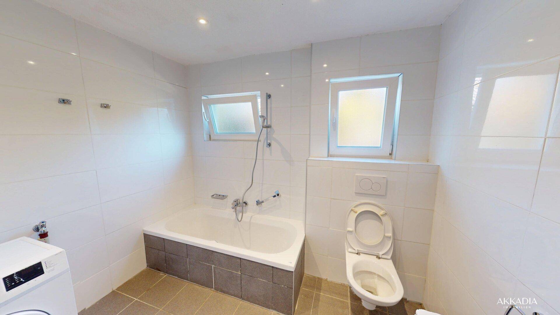 Modernes Badezimmer mit Badewanne, Toilette und zwei Fenstern für Tageslicht.