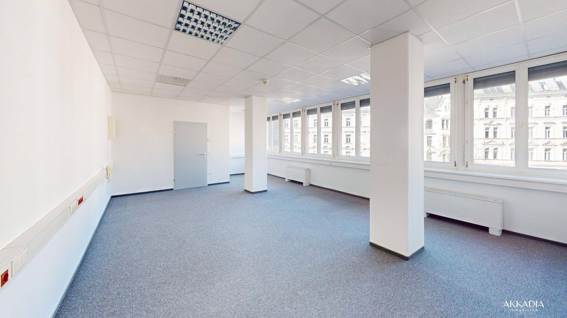 Leerer Büroraum mit Teppichboden, weißen Wänden und einer Reihe von Fenstern mit Stadtblick.