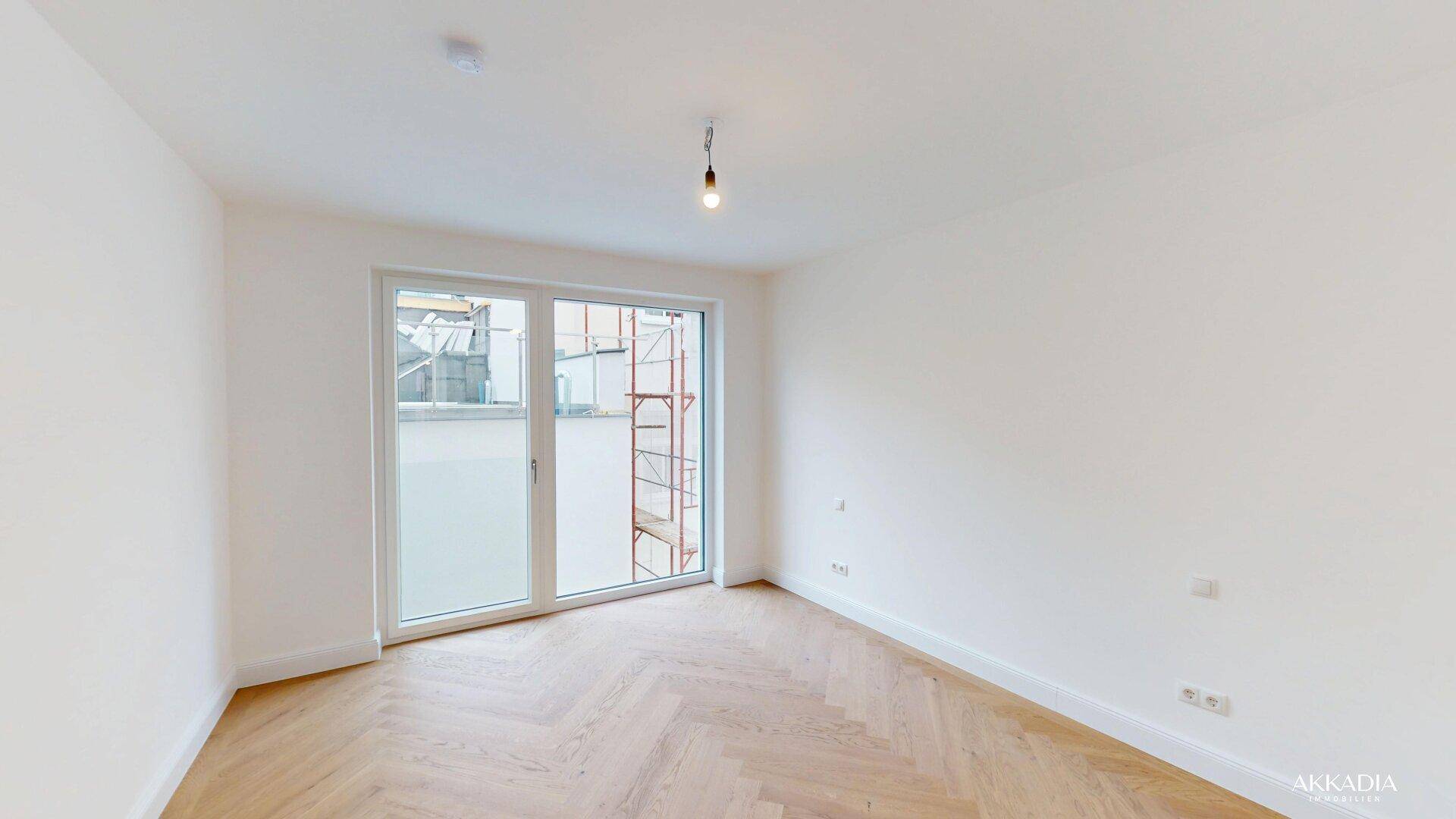 Heller Raum mit Fischgrätparkett und großer Fenstertür, die viel Tageslicht hereinlässt.