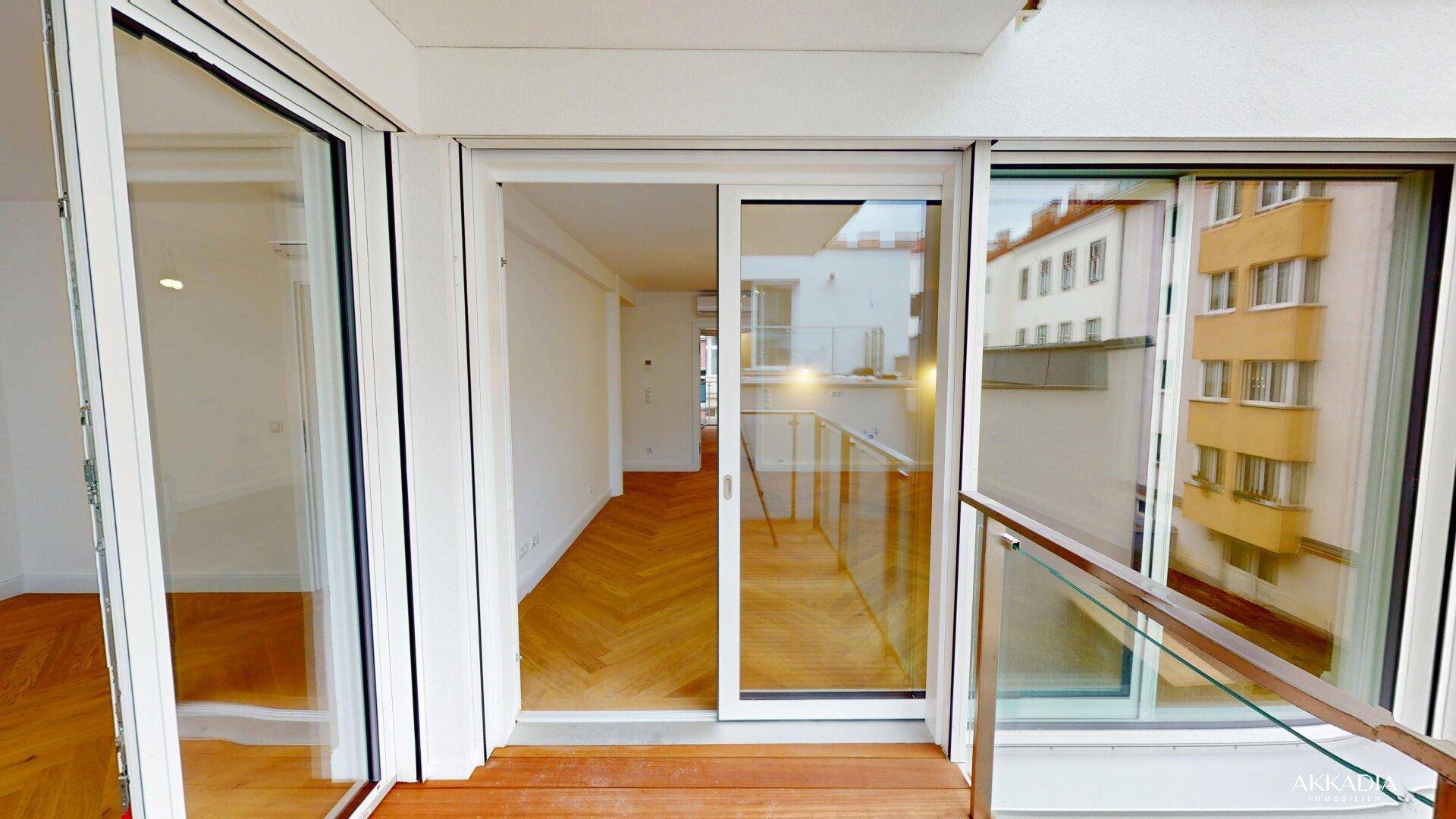 Balkon mit Holzboden und Glasgeländer, der in einen hellen Innenraum mit Fischgrätparkett führt.