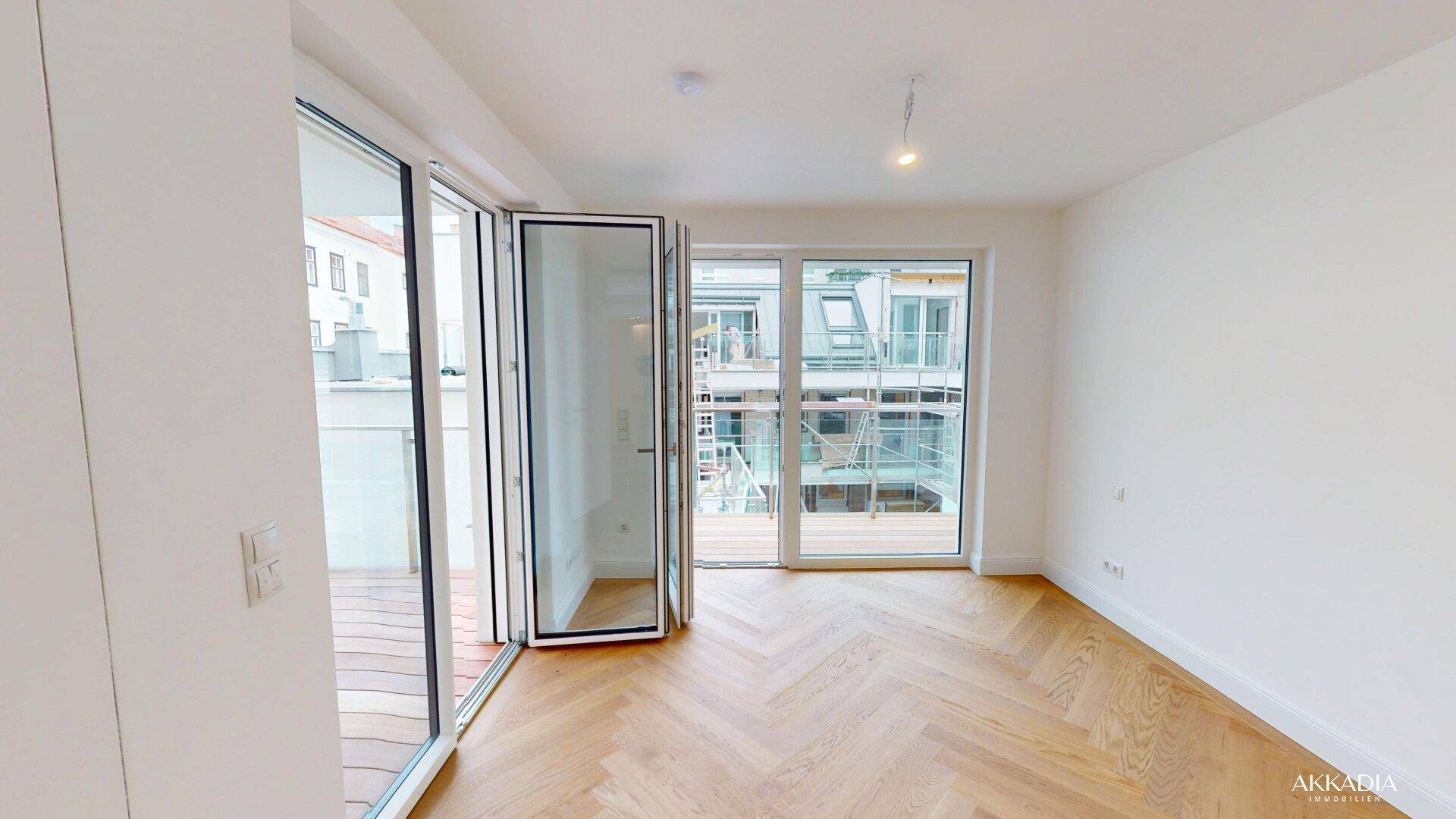 Lichtdurchflutetes Zimmer mit Fischgrätparkett und großen Fenstern zum Balkon mit Stadtblick.