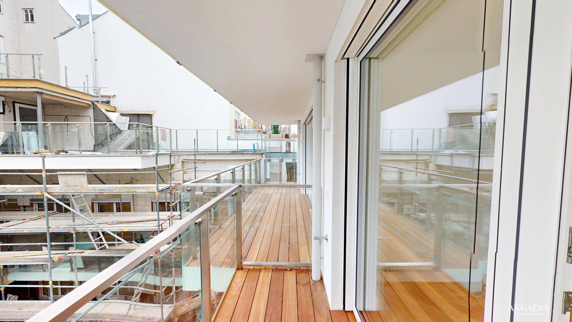 Langer Balkon mit Holzboden und Glasgeländer, bietet Blick auf eine Baustelle und umliegende Gebäude.
