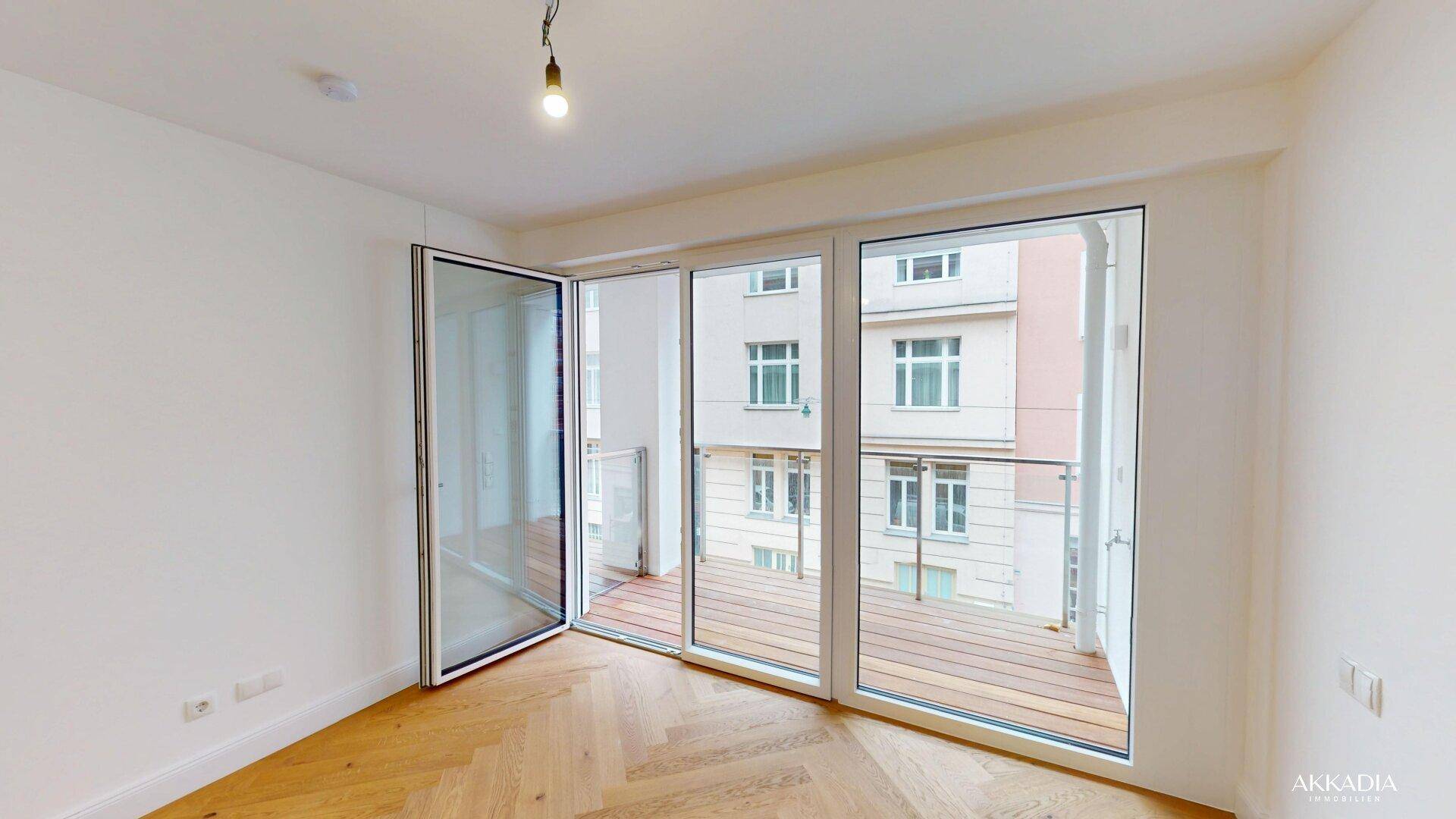 Heller Raum mit Fischgrätparkett und großen Fenstern, die auf einen Balkon führen.