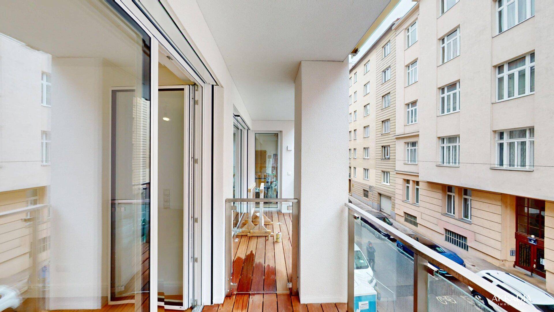 Langer Balkon mit Holzboden und Glasgeländer, bietet Blick auf die Straße und umliegende Gebäude.