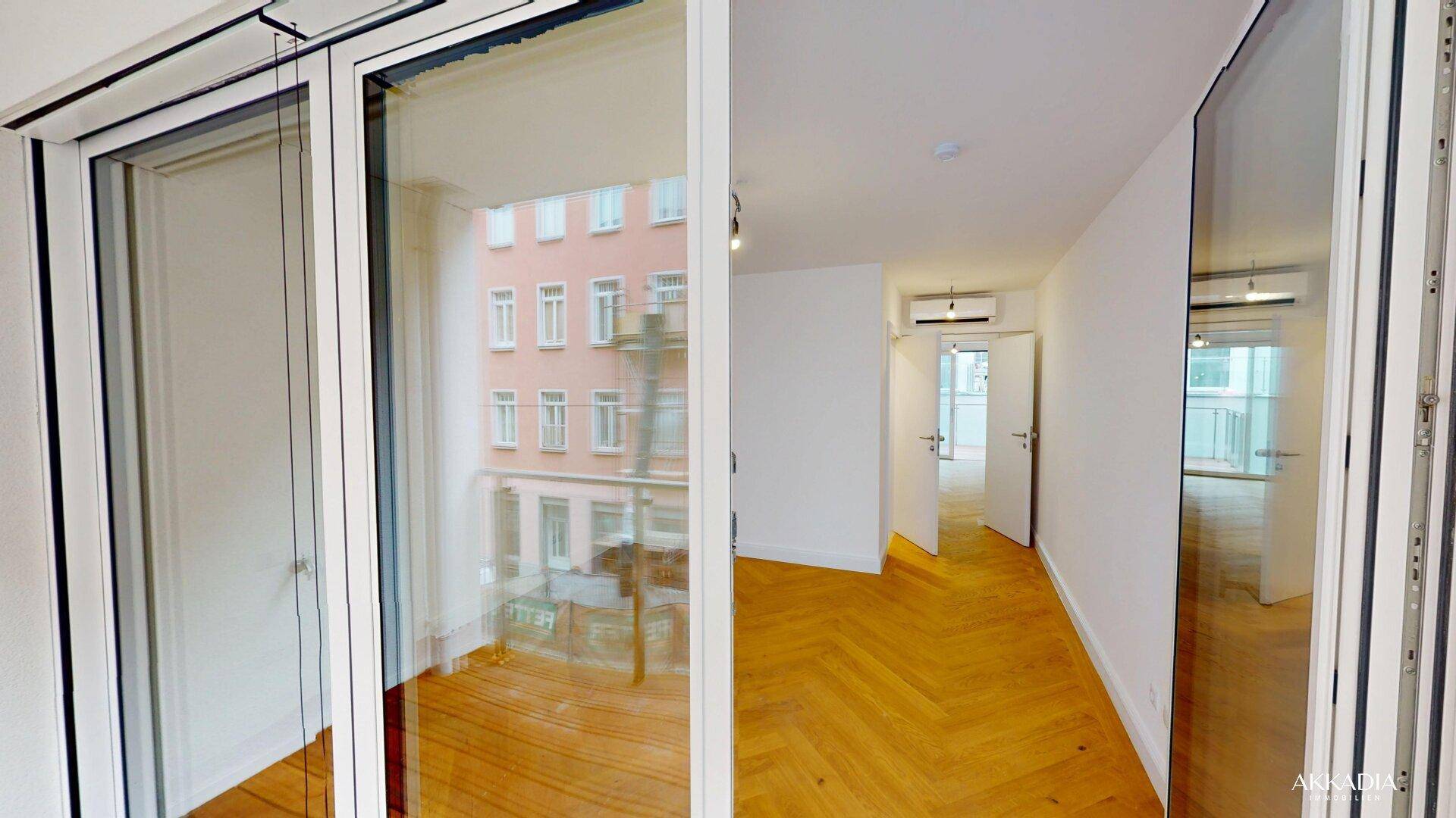 Balkon mit Holzboden und Glasgeländer, der in einen hellen Innenraum mit Fischgrätparkett führt.