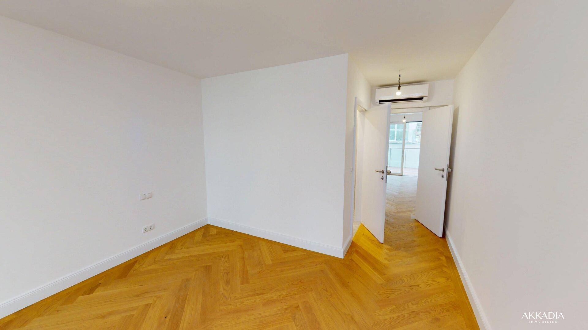 Geräumiger, unmöblierter Raum mit hellem Fischgrätparkett und Klimaanlage an der Wand.